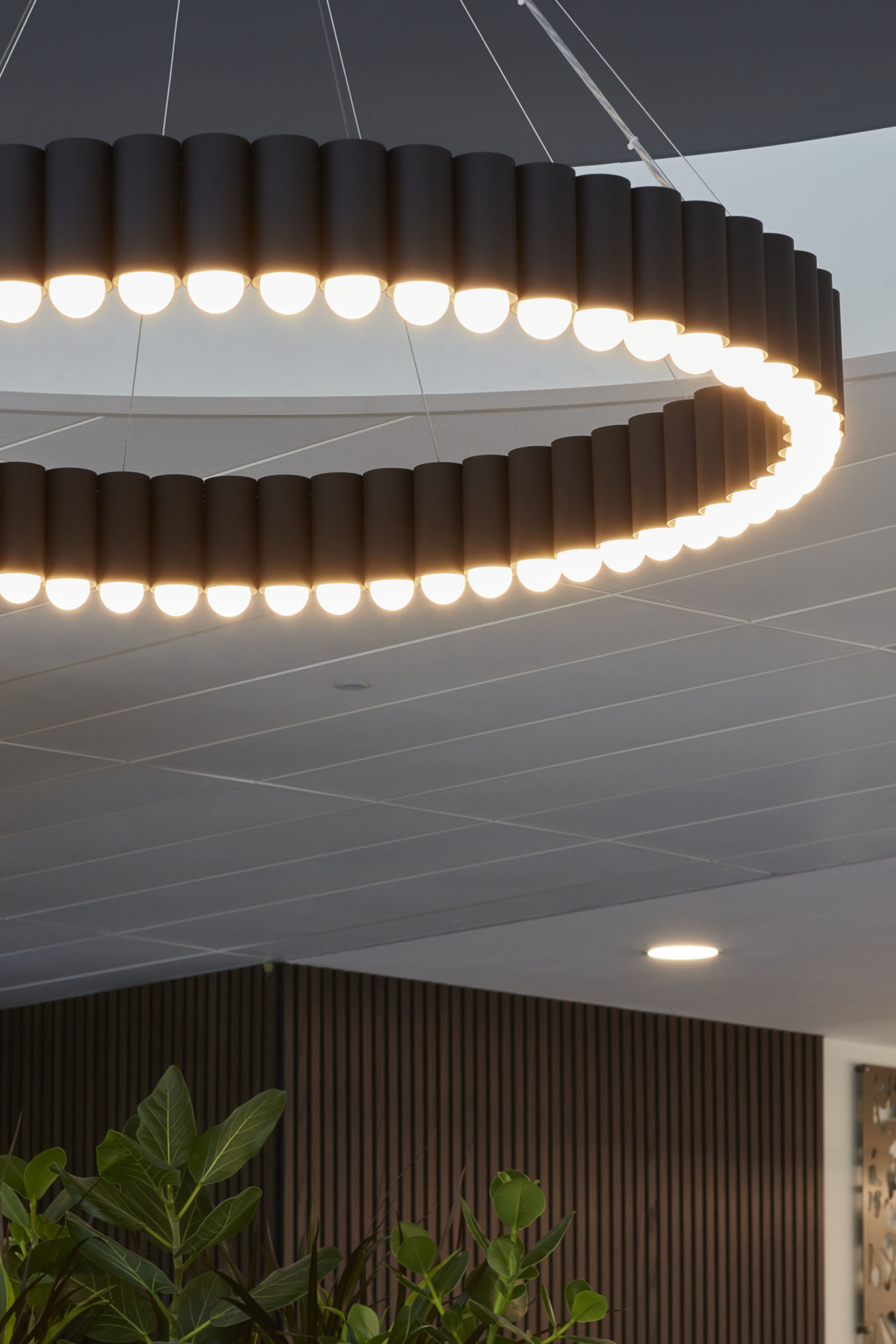 A circular chandelier emits light, suspended from a ceiling in a modern interior with plants and decorative wood panels.
