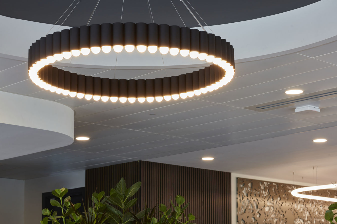 A circular chandelier emits light, suspended from a ceiling in a modern interior with plants and decorative wood panels.