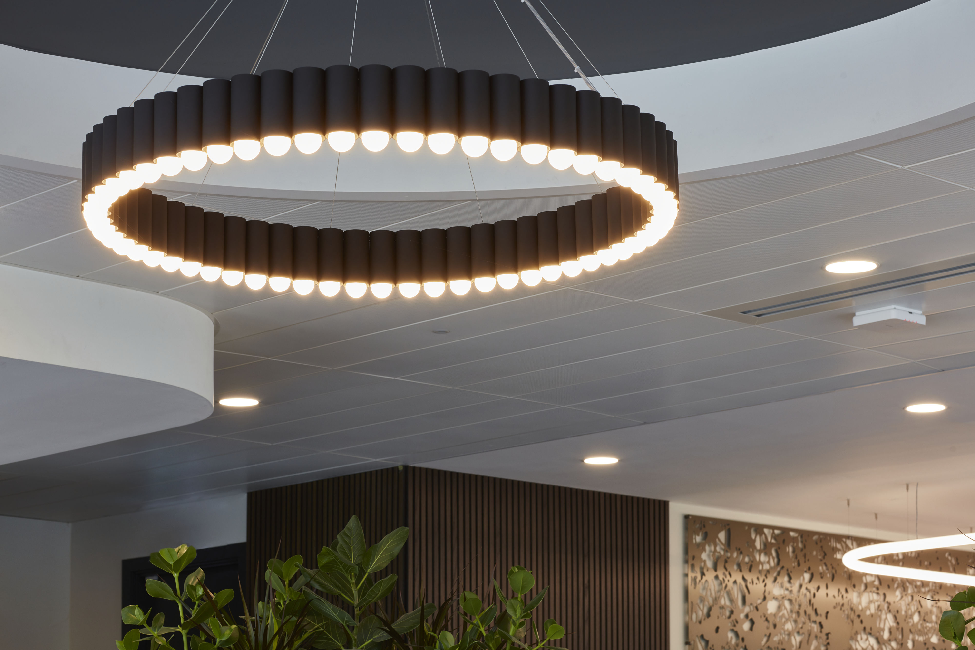A circular chandelier emits light, suspended from a ceiling in a modern interior with plants and decorative wood panels.