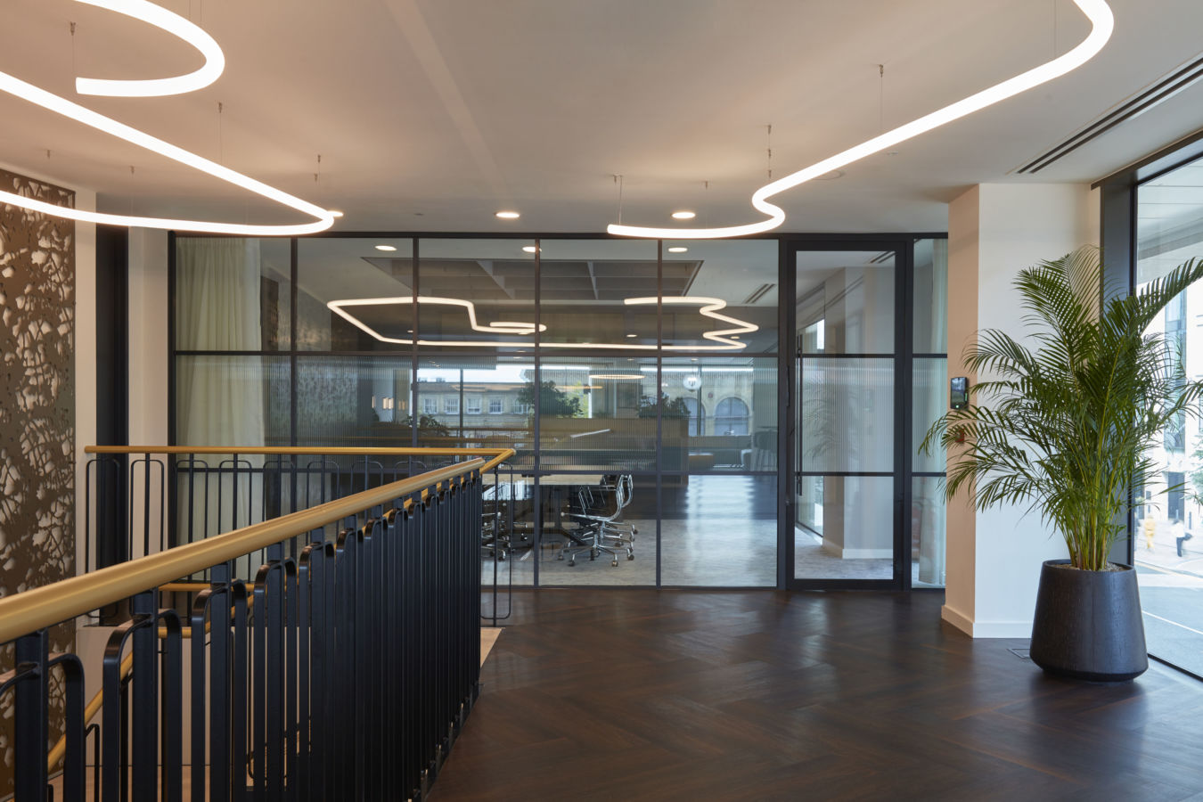 Office interior features modern lighting fixtures illuminating a glass-walled conference room and a wooden staircase. A large potted plant decorates the entrance, enhancing the contemporary design.