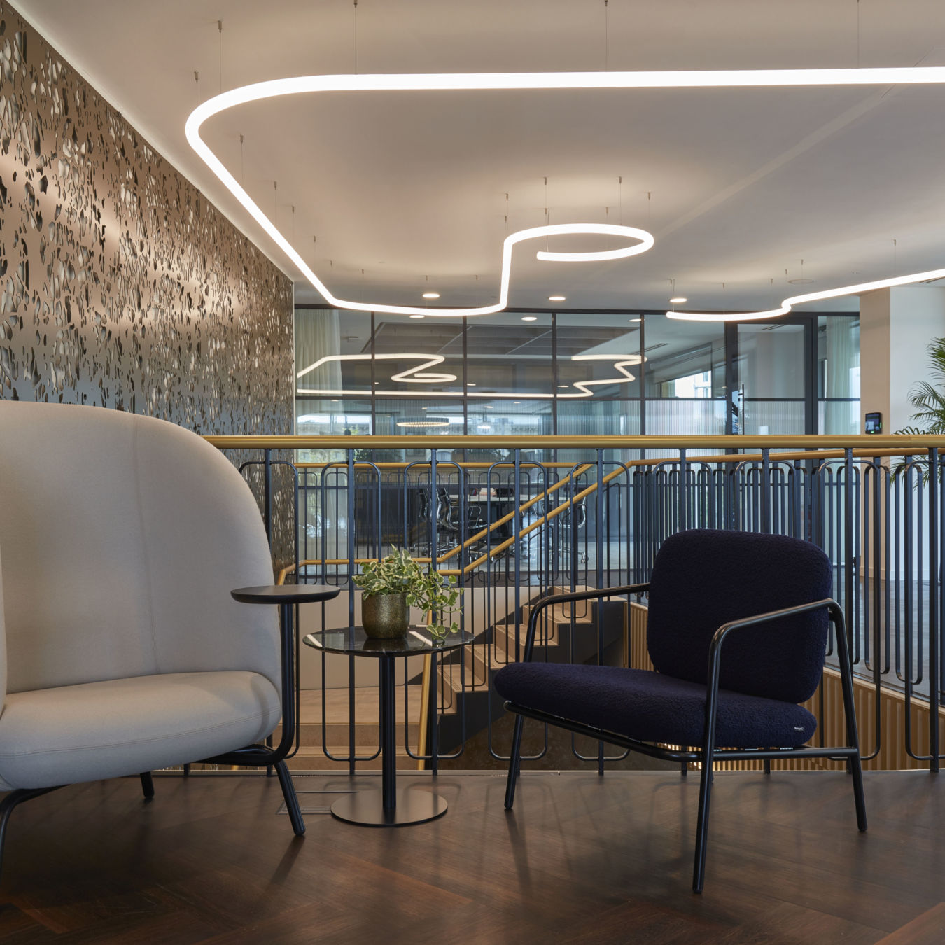 A modern office lounge features two stylish chairs surrounding a small table with a plant, set against a decorative wall and glass-walled meeting rooms, with overhead curved lighting fixtures.