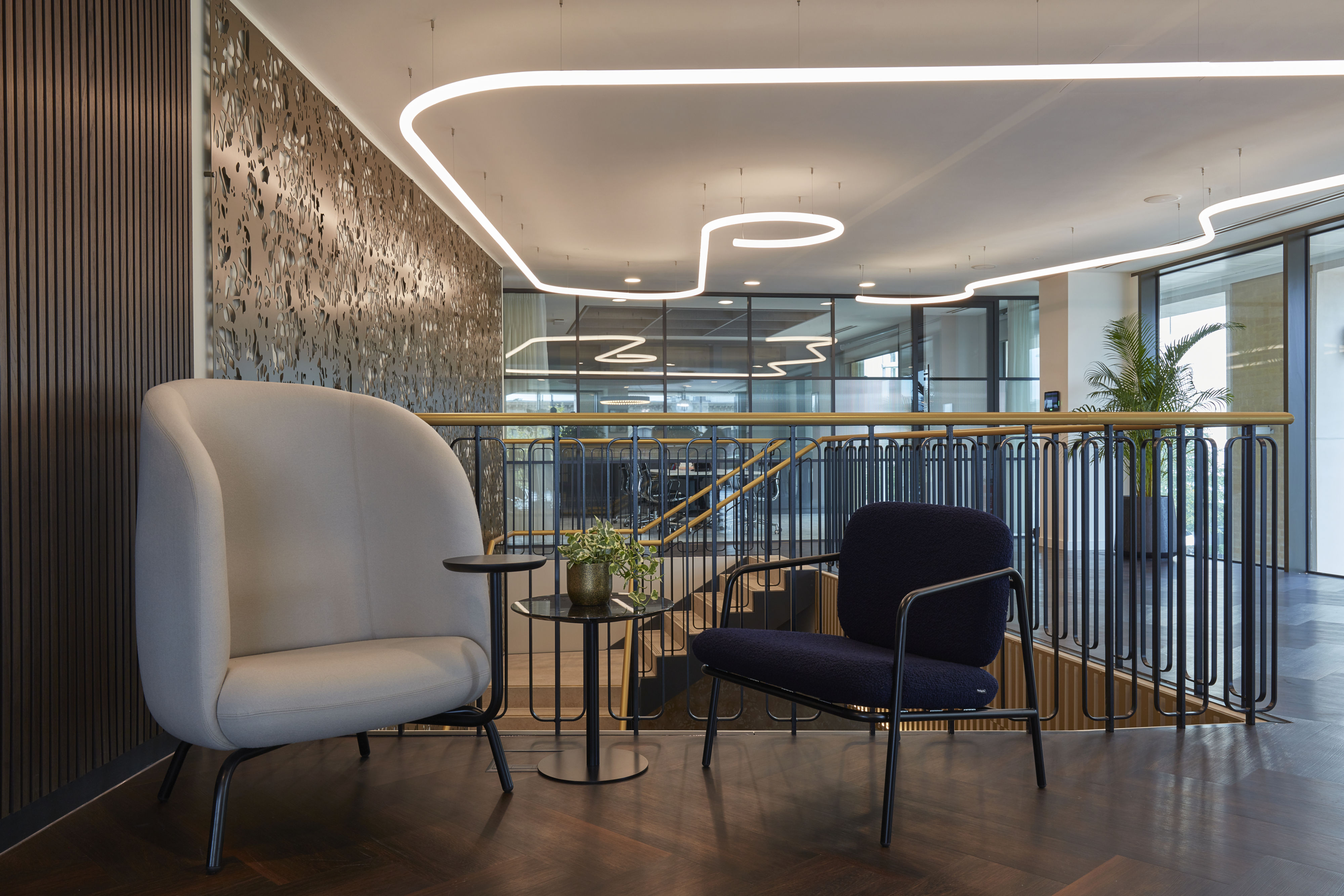 A modern office lounge features two stylish chairs surrounding a small table with a plant, set against a decorative wall and glass-walled meeting rooms, with overhead curved lighting fixtures.