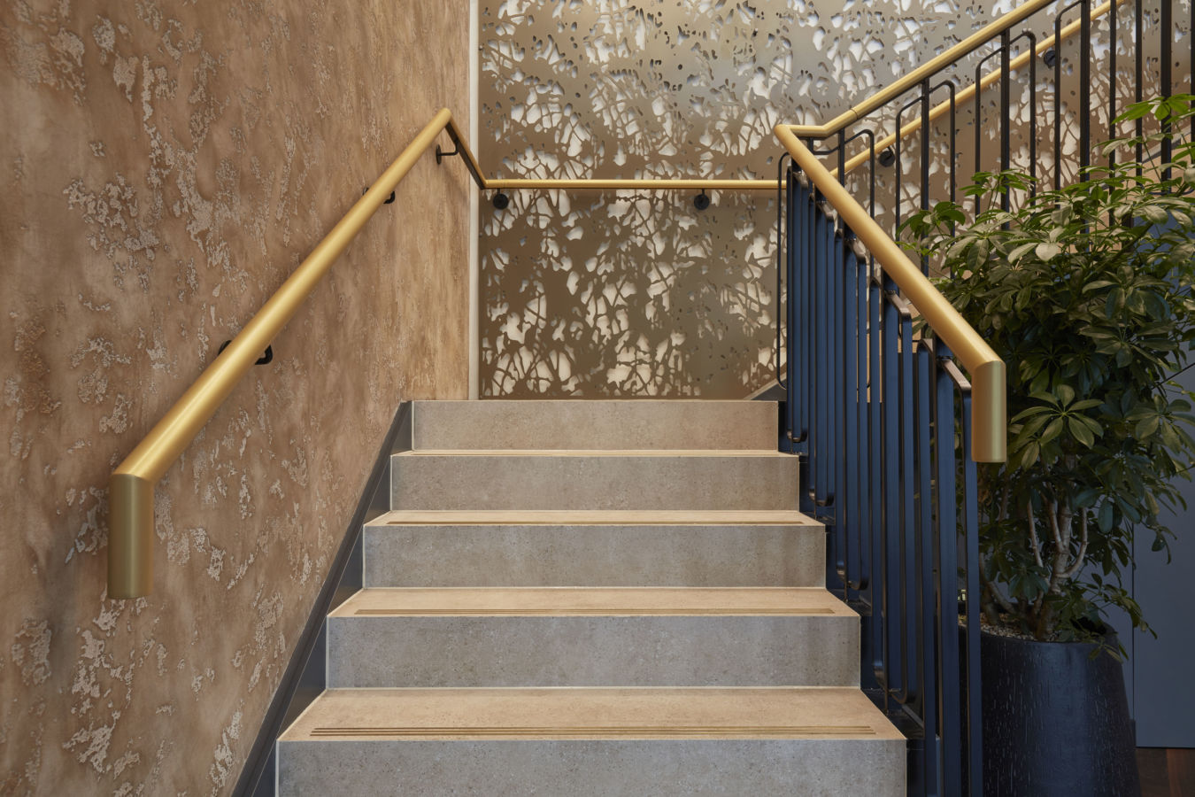 Staircase features beige steps ascending toward a decorative wall with an abstract, cut-out pattern, surrounded by gold railings, creating a modern, artistic interior design environment.