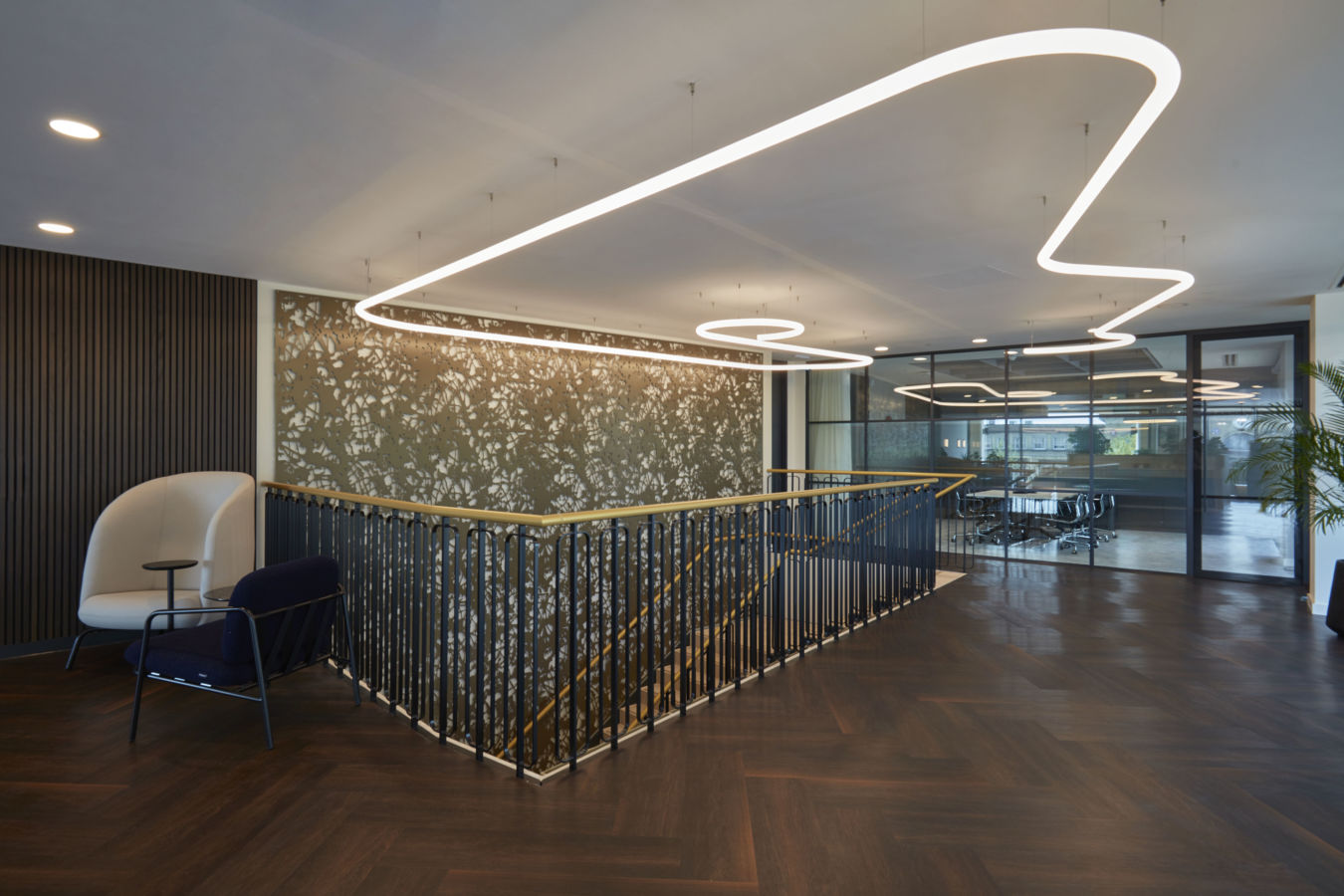 Modern office interior features sleek overhead lighting forming abstract shapes, with stylish furnishings and dark wood flooring, surrounding a central staircase near glass-walled meeting rooms and decorative wall panels.
