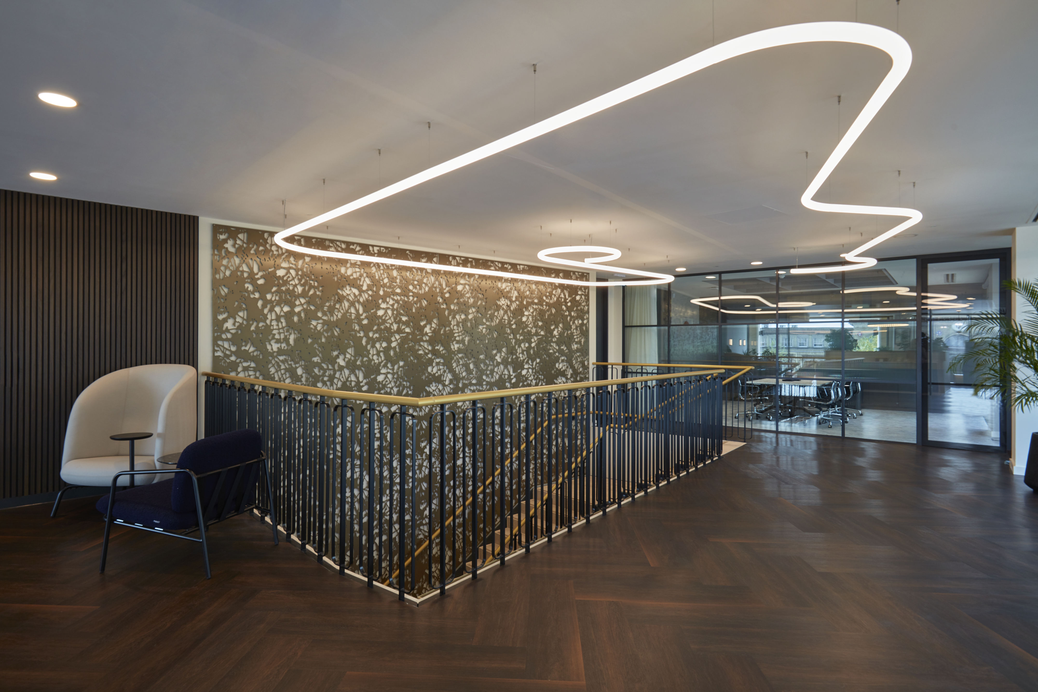 Modern office interior features sleek overhead lighting forming abstract shapes, with stylish furnishings and dark wood flooring, surrounding a central staircase near glass-walled meeting rooms and decorative wall panels.