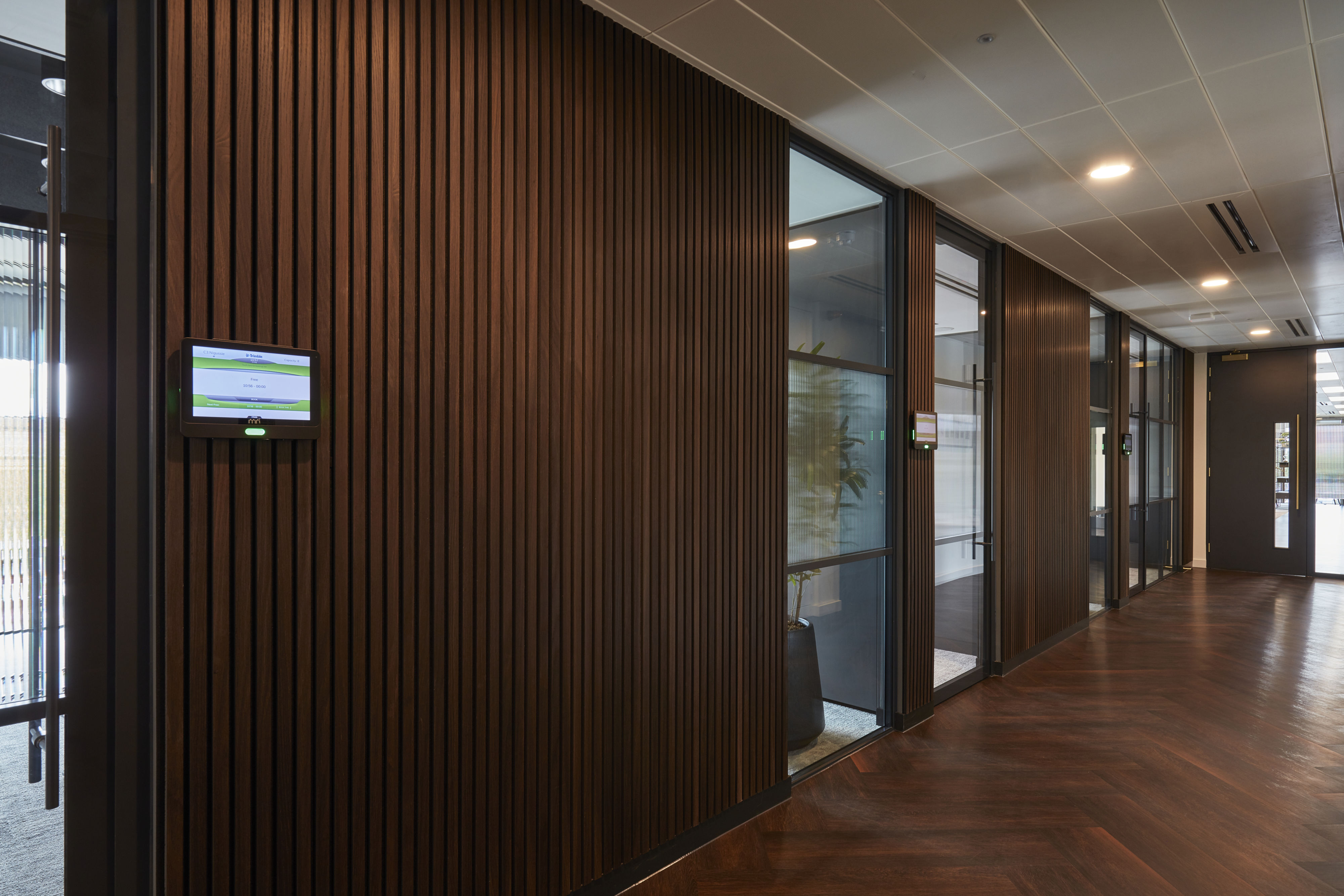 Wood-paneled hallway featuring a digital screen displaying meeting room information; glass doors on the right reveal an office setting with plants, illuminated by overhead lights.