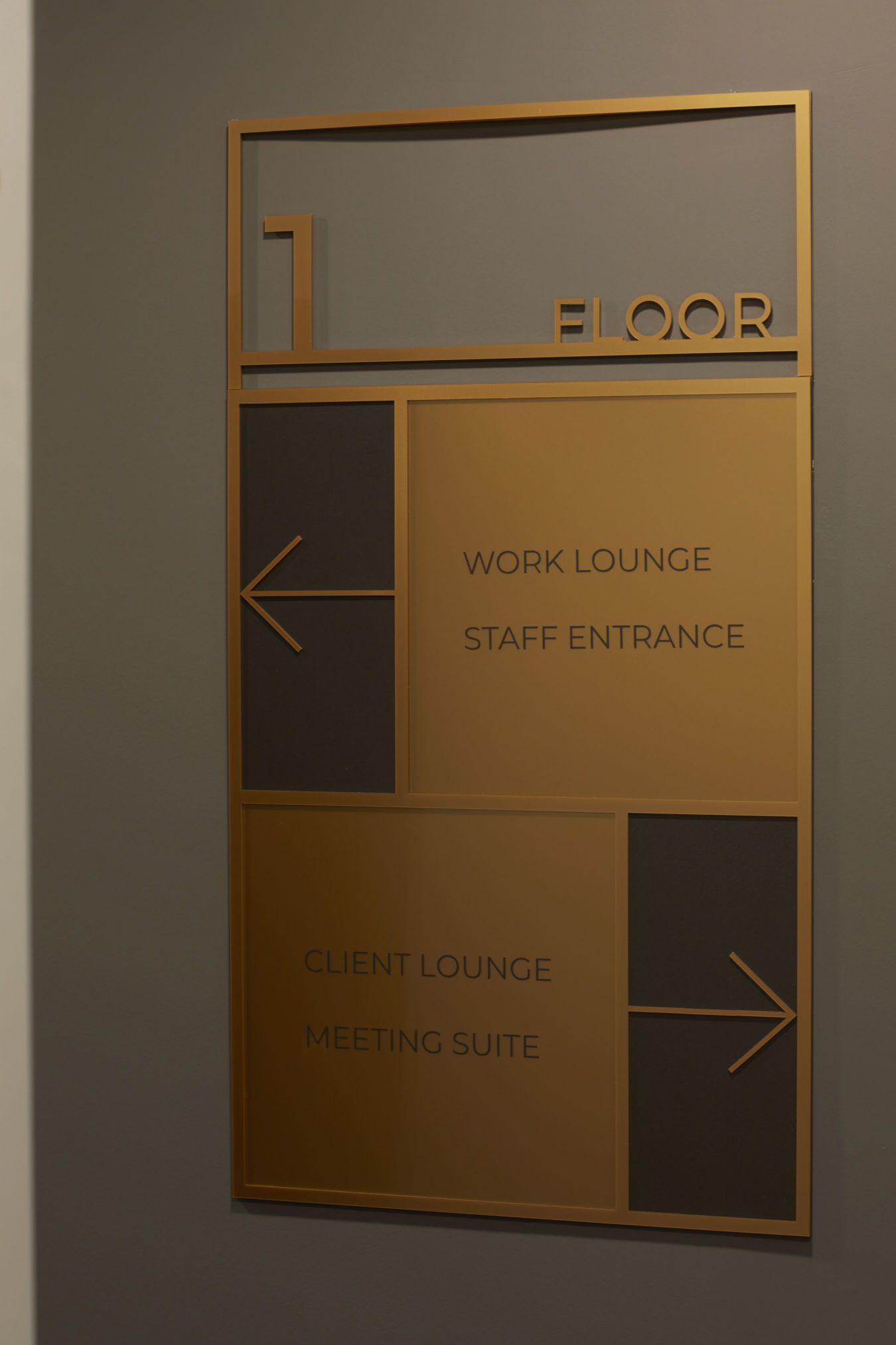 Signage displaying “1 FLOOR” with directional arrows. Left arrow: “WORK LOUNGE,” “STAFF ENTRANCE.” Right arrow: “CLIENT LOUNGE,” “MEETING SUITE.” Set against a gray wall.