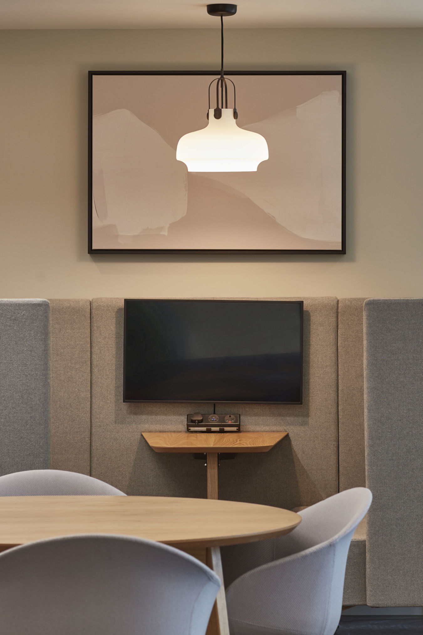 A round wooden table with chairs sits in front of a wall-mounted TV, surrounded by gray acoustic panels, under a framed abstract art piece and modern light fixture, with indoor plants nearby.