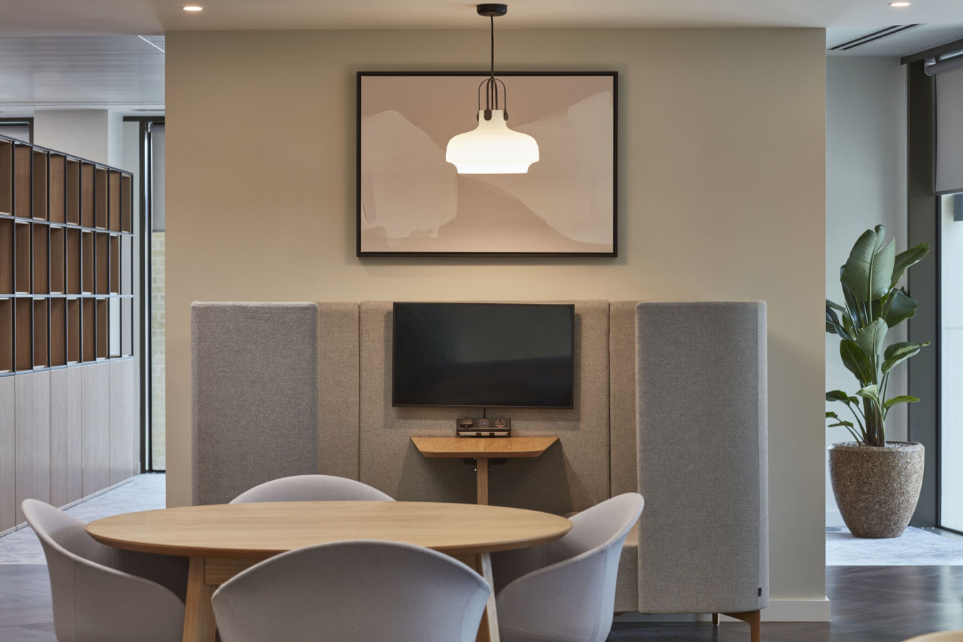 A round wooden table with chairs sits in front of a wall-mounted TV, surrounded by gray acoustic panels, under a framed abstract art piece and modern light fixture, with indoor plants nearby.