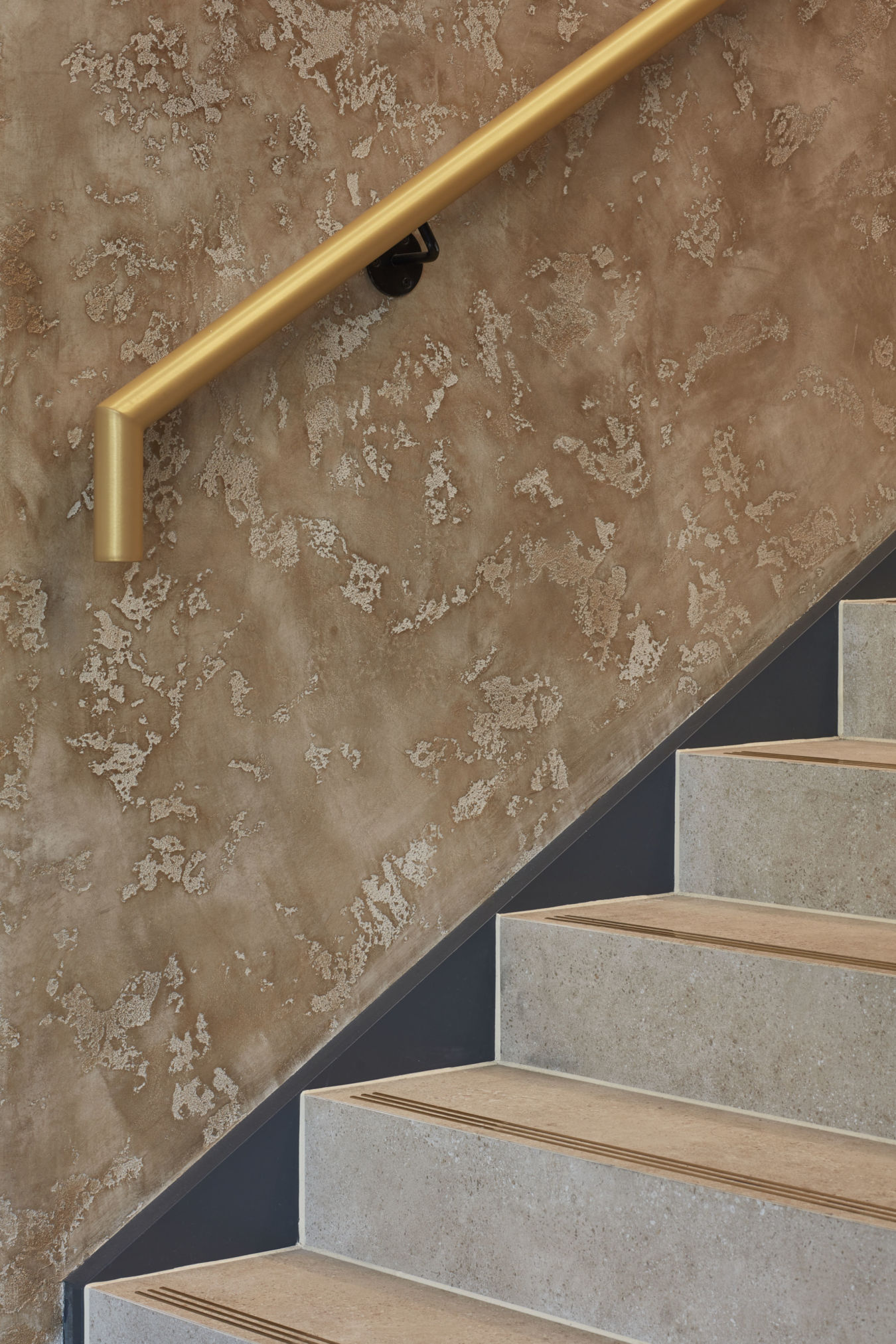 Golden handrail angles above textured beige wall, complementing adjacent light brown stairs with subtle tread markings in a contemporary interior setting.