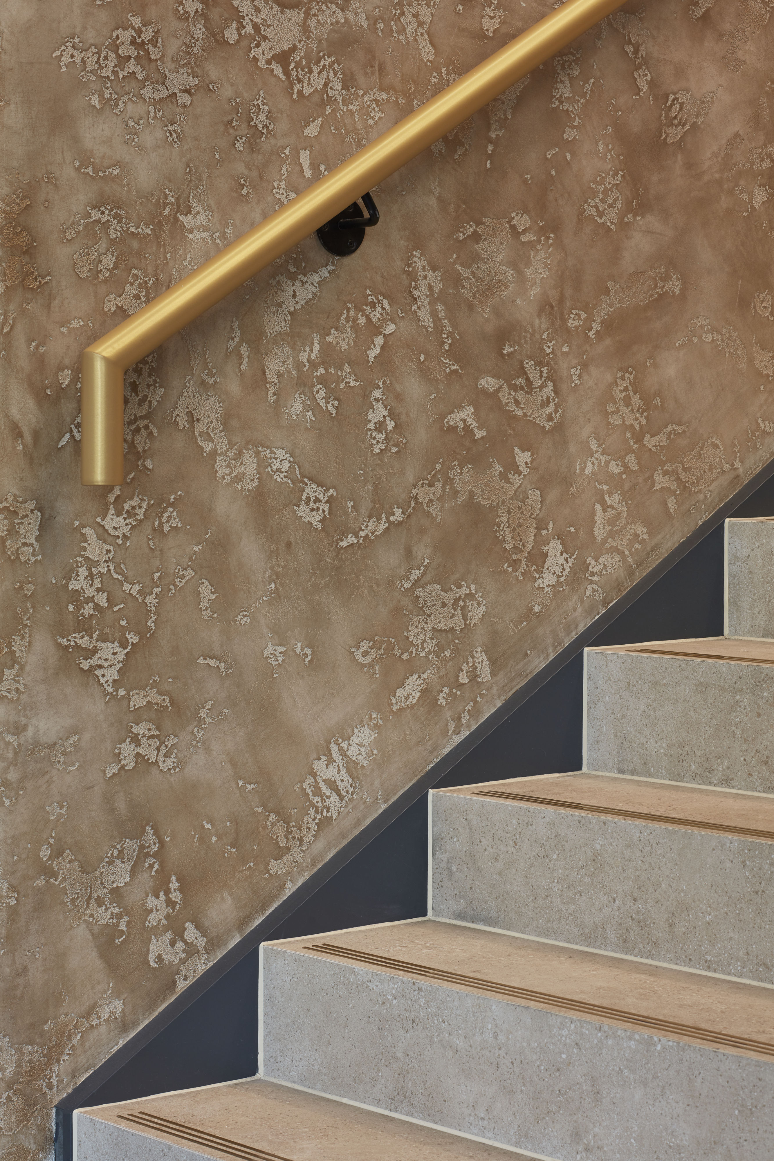Golden handrail angles above textured beige wall, complementing adjacent light brown stairs with subtle tread markings in a contemporary interior setting.