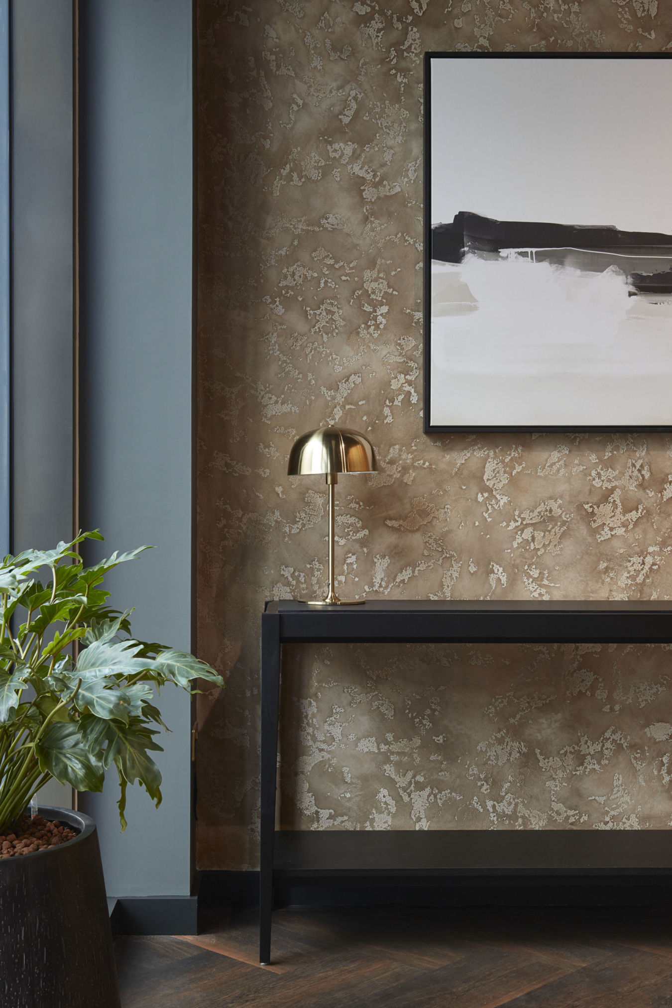 A modern interior scene features a large potted plant by a frosted window, a black table with a golden lamp, and minimalist decor against a textured brown wall with abstract artwork.