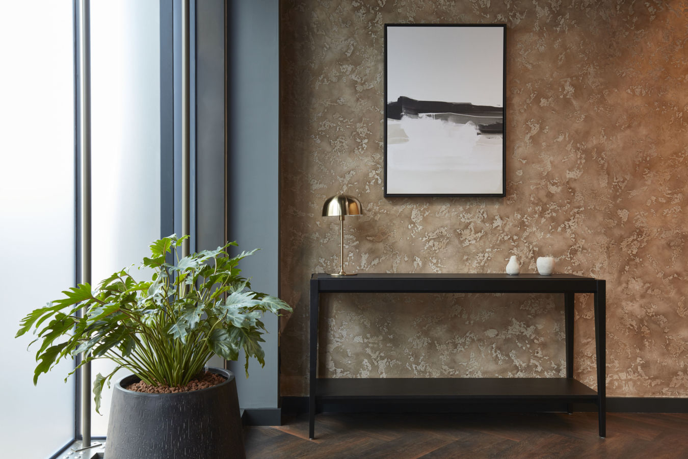 A modern interior scene features a large potted plant by a frosted window, a black table with a golden lamp, and minimalist decor against a textured brown wall with abstract artwork.