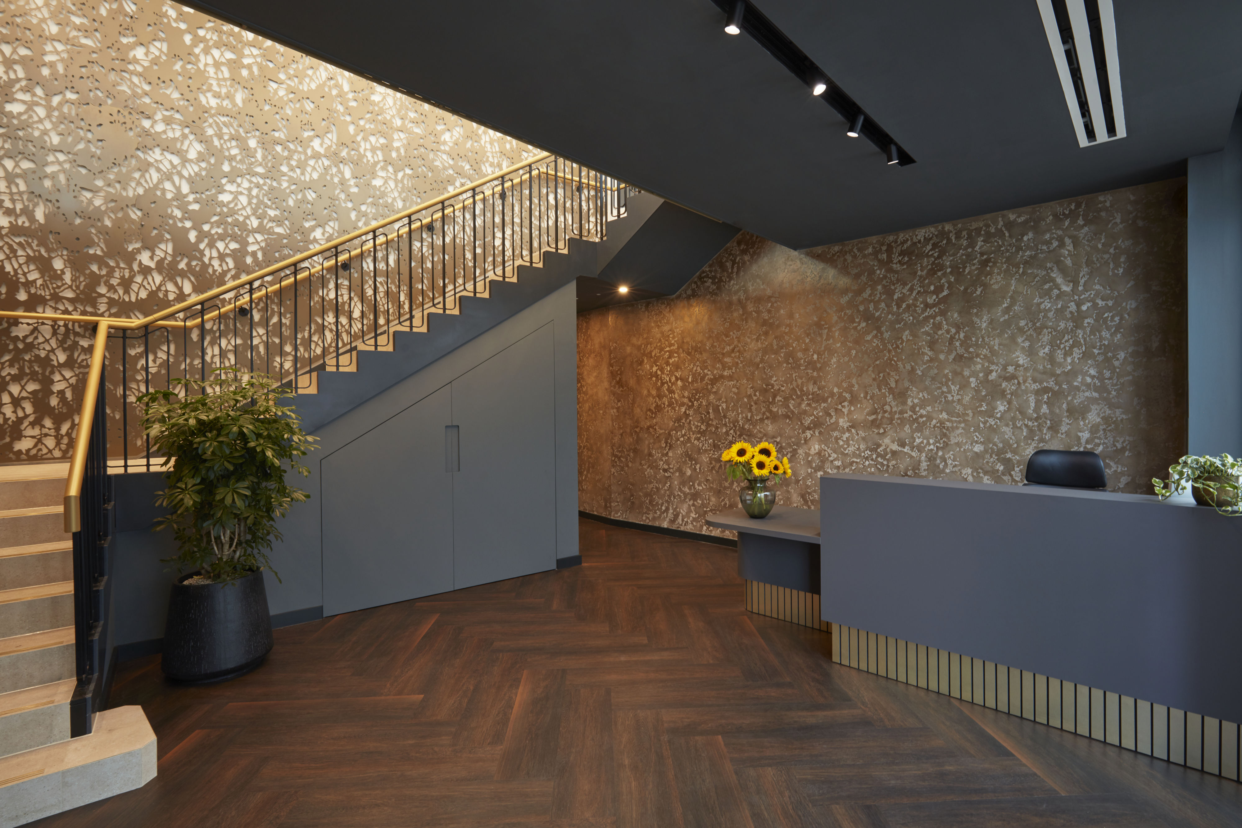 A modern reception area features a blue desk with flowers, a patterned wall, and a staircase with plants, creating a warm and inviting atmosphere.