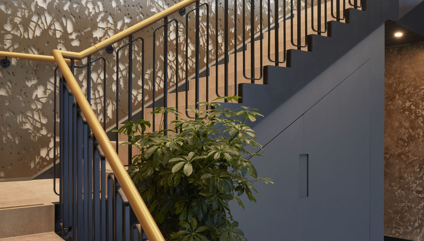 Staircase with beige steps and gold handrail ascends beside a decorative perforated wall with organic patterns; a potted plant sits at the bottom on dark wood flooring.
