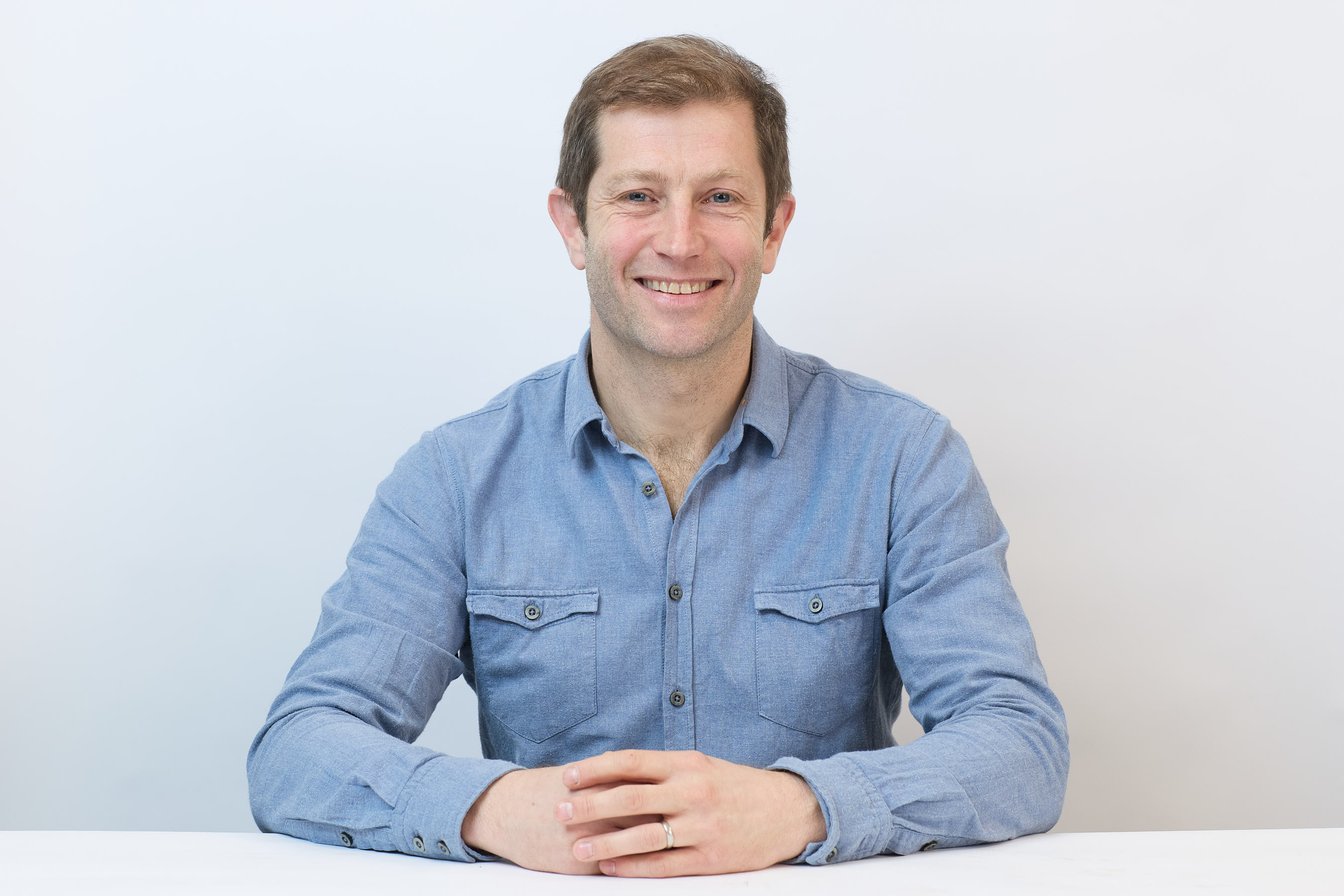 Smiling man in a blue shirt sits with hands clasped on a white table, against a plain light background.