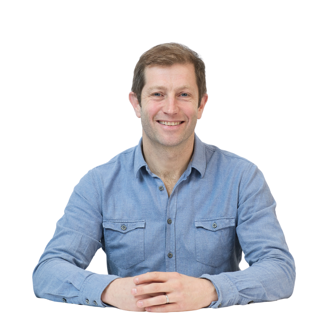 A person wearing a blue button-up shirt smiles while sitting at a table with hands folded, isolated on a plain white background.