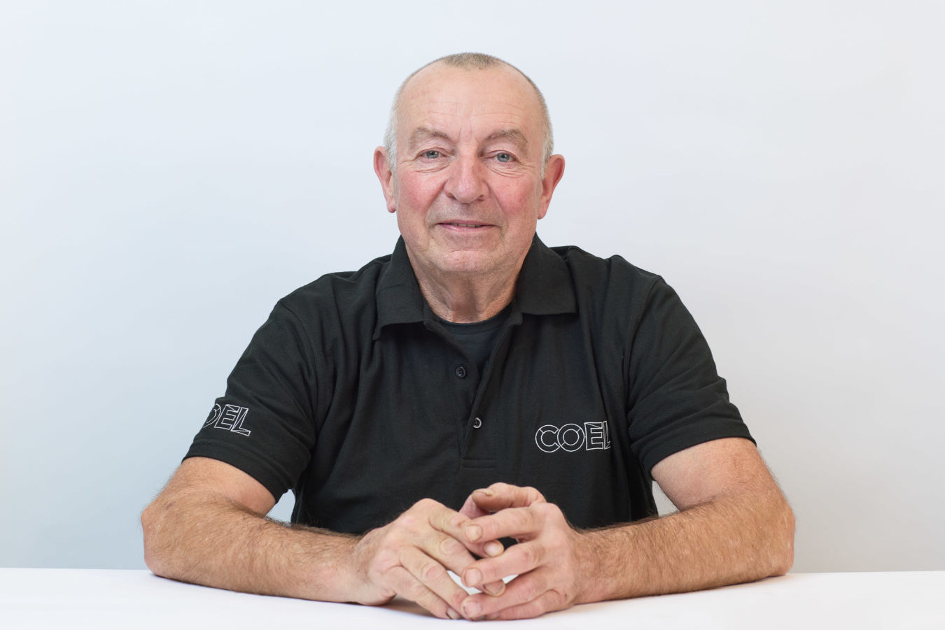 A man wearing a black polo shirt with "COEL" text, sitting with hands clasped, against a plain white background.