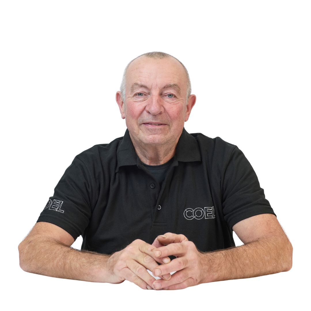 An older man wearing a black polo shirt with the text "COEL" sits with hands clasped, looking directly at the camera against a white background.