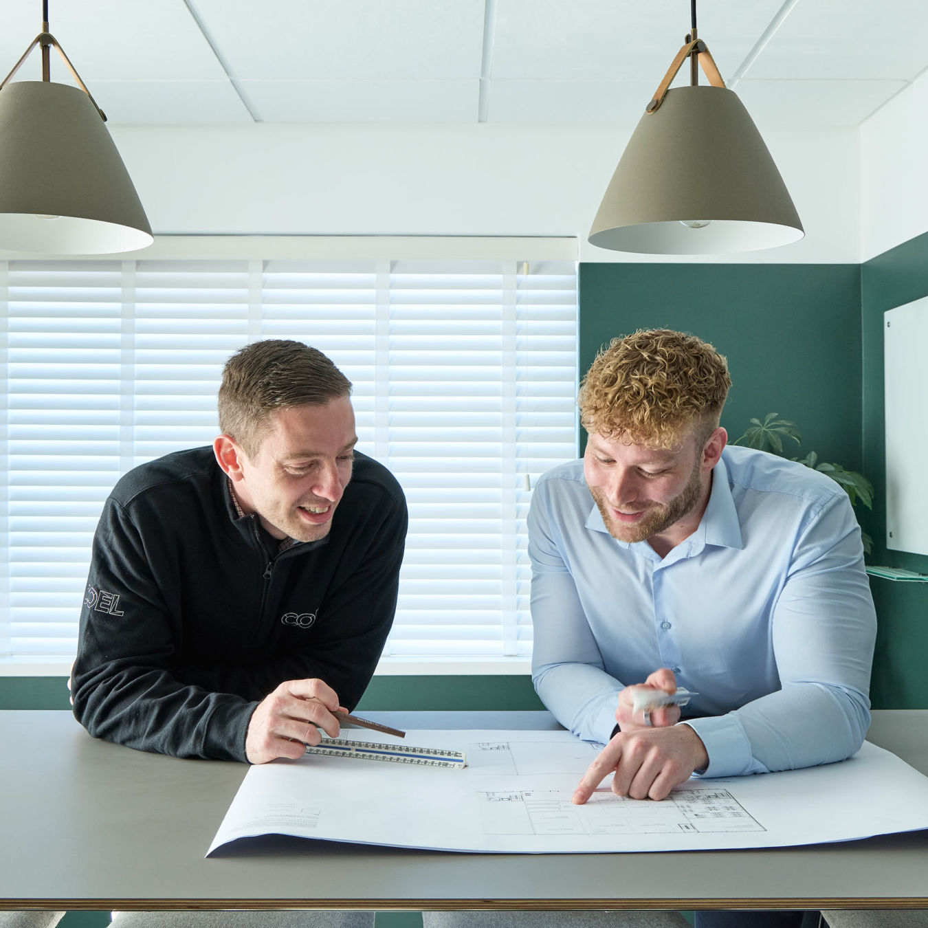 Two men examine architectural blueprints on a table, smiling and discussing, in a bright office with white blinds and green walls. One man wears a black jacket with a logo.