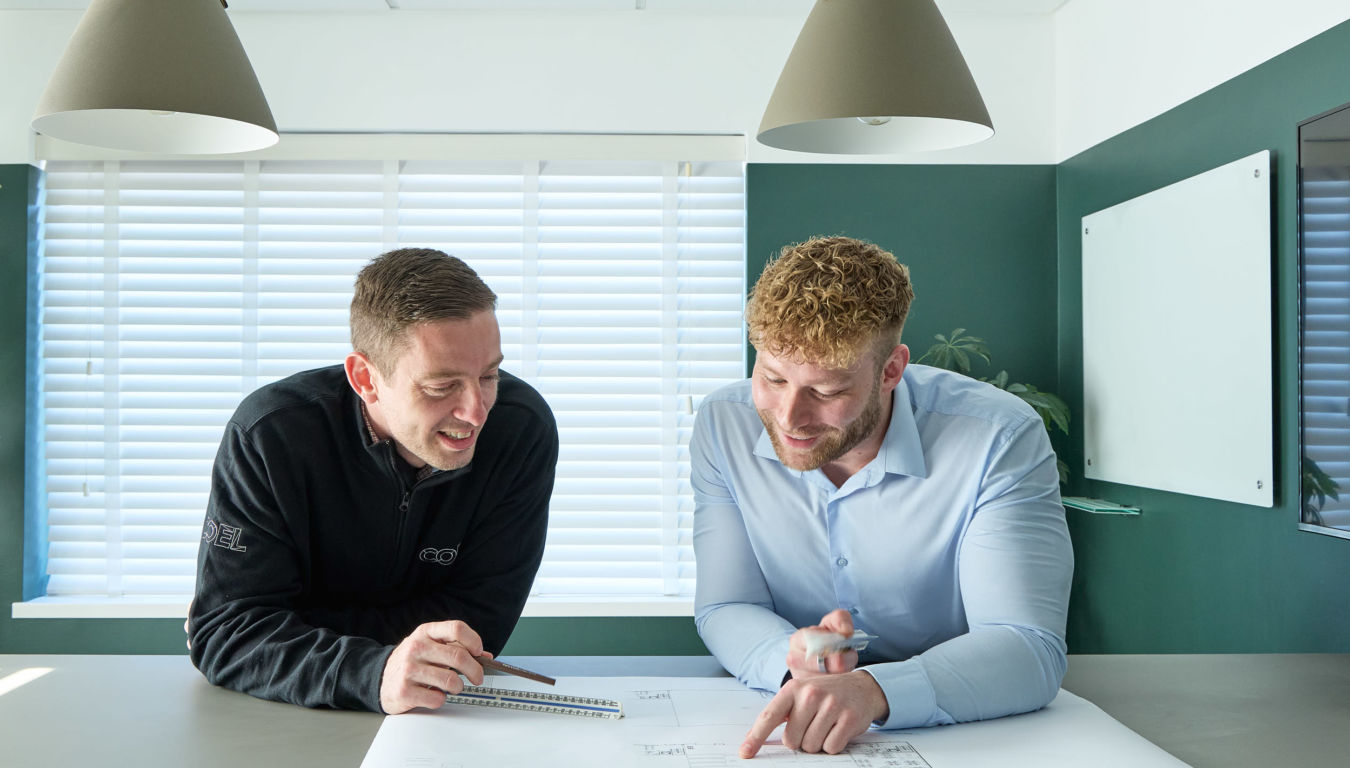 Two men discuss a blueprint on a table in a modern office with pendant lights, a whiteboard, and large windows with blinds. One wears a black jacket with "COEL" text.