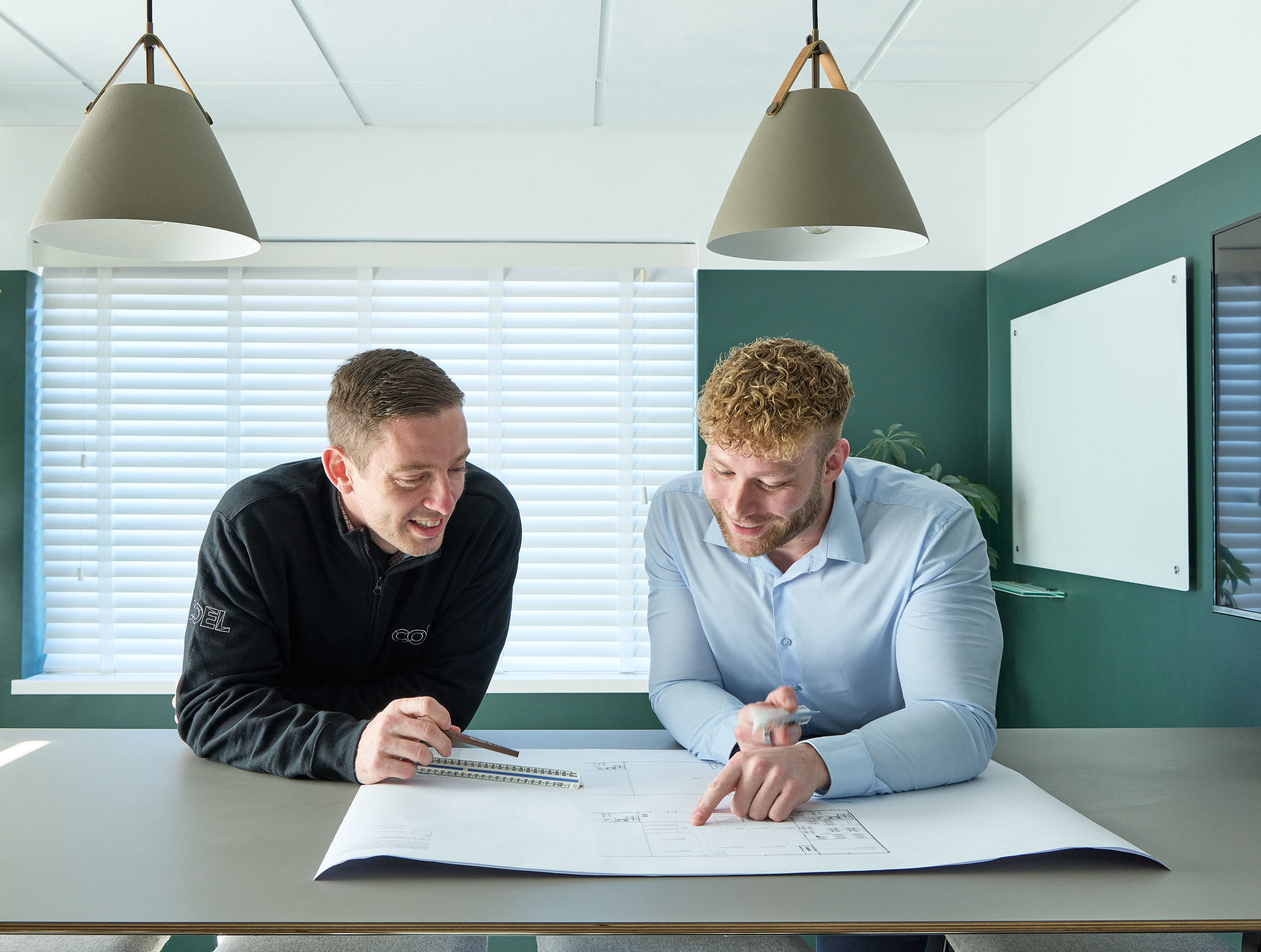 Two men discuss a blueprint on a table in a modern office with pendant lights, a whiteboard, and large windows with blinds. One wears a black jacket with "COEL" text.