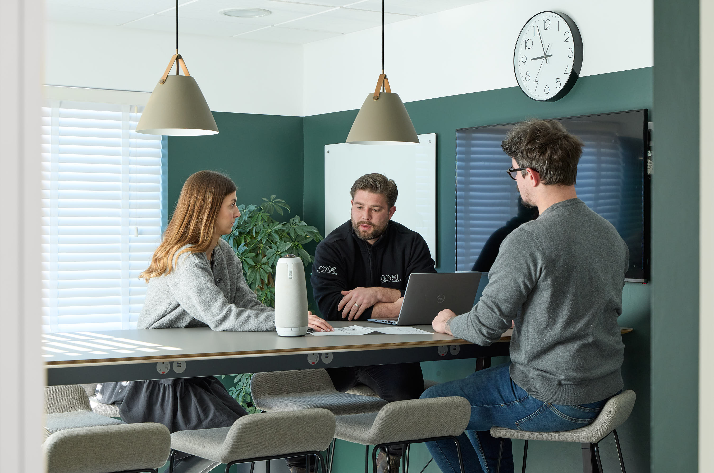 Three people discuss at a conference table with a laptop and speaker. Green walls, a wall clock showing 10:09, and pendant lighting create an office setting.