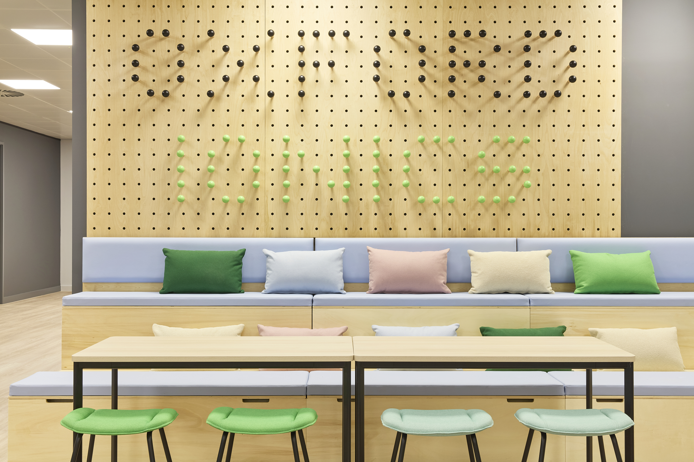 Wooden pegboard wall with text "SWIFT MADE MOVICS" in black and green balls above cushioned seating with colorful pillows; tables and green stools in a modern, bright room.