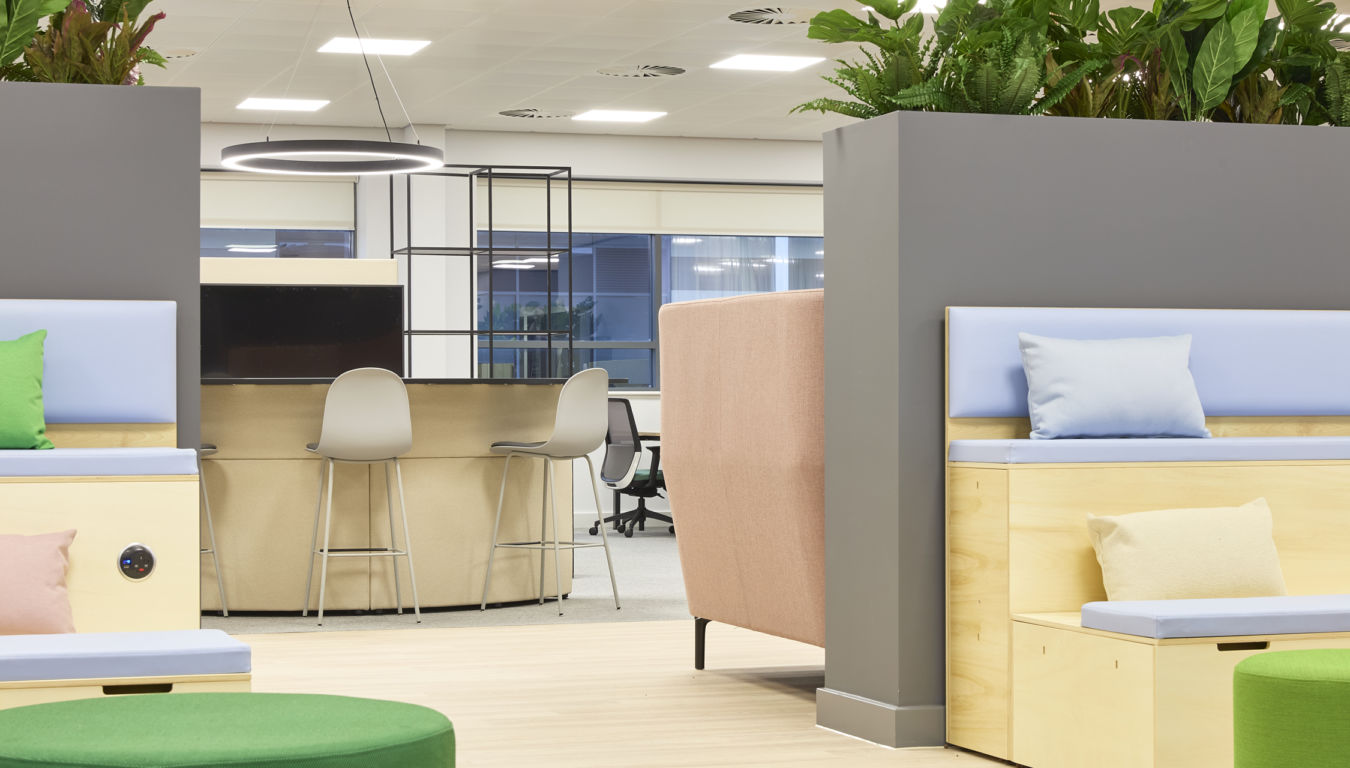 Open-plan office with mixed seating, high counter stools, soft furnishings, indoor plants, and modern lighting