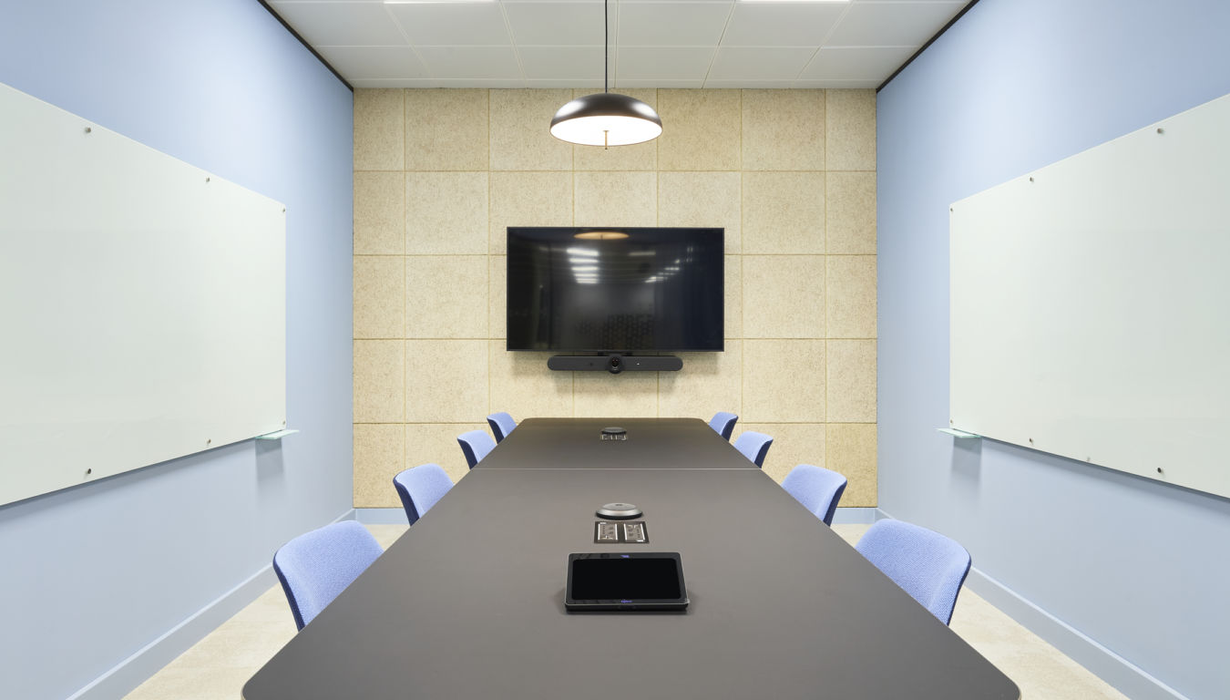 Conference room featuring a long black table with eight blue chairs, centered under a pendant lamp. A large screen and conferencing device are mounted on a beige panel wall.