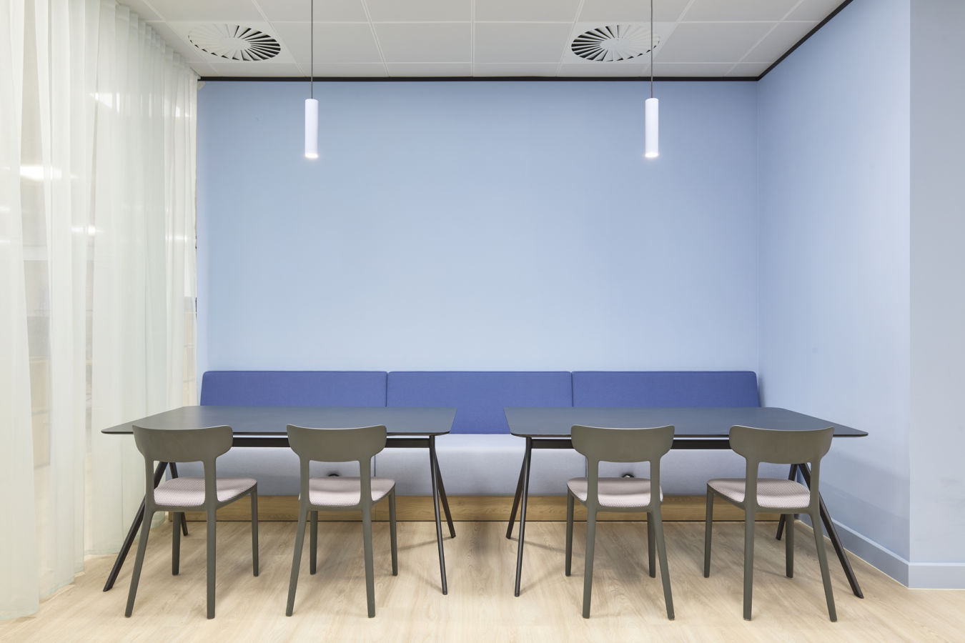 A modern meeting space with light blue walls, two black tables, six gray chairs, a blue bench, and two pendant lights, creating a calm atmosphere