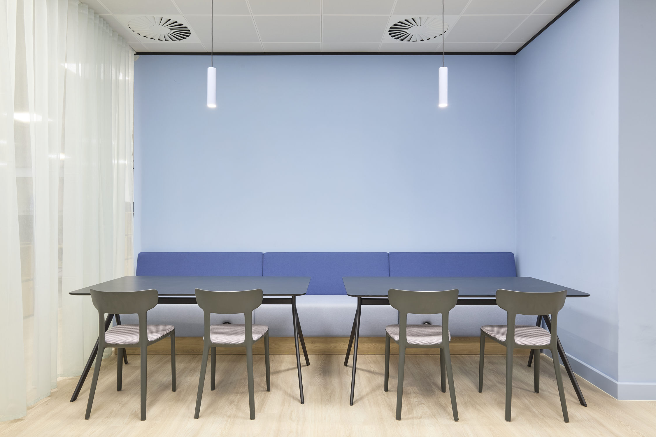 A modern meeting space with light blue walls, two black tables, six gray chairs, a blue bench, and two pendant lights, creating a calm atmosphere