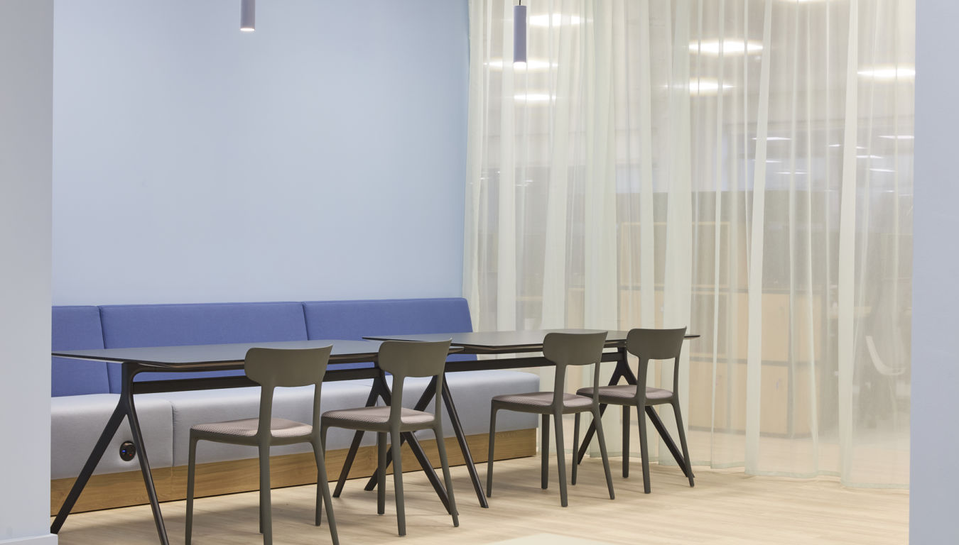 A modern office space features a row of chairs and tables against a blue wall, accompanied by soft lighting and sheer curtains in the background.