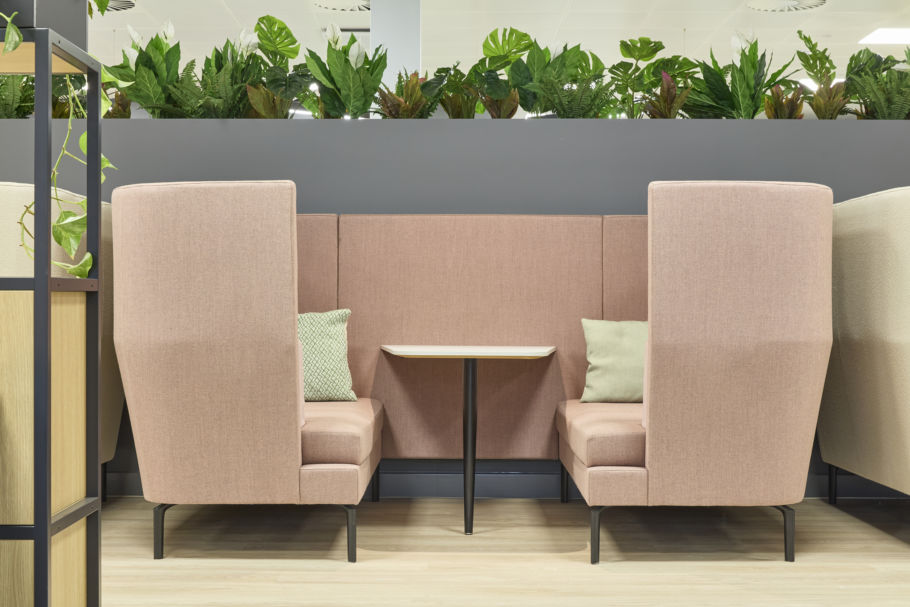 cozy seating area features two soft pink high-backed booths facing each other with a small table between. Green plants line the top of a gray wall, adding a fresh touch