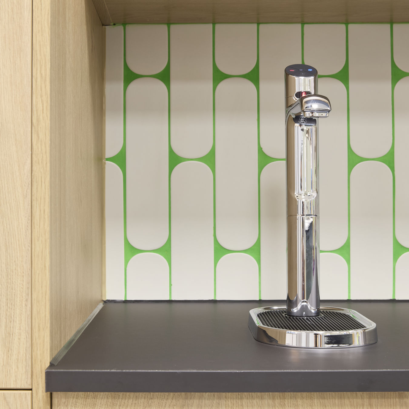 A chrome water dispenser stands on a black countertop, set against a patterned green and white tiled backsplash in a wooden kitchen cabinet area.