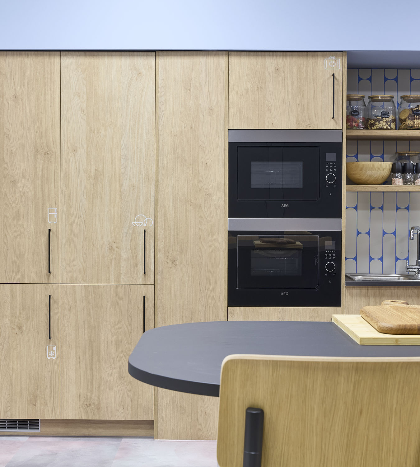 Modern kitchen with light wood cabinets and dual AEG ovens, against a geometric blue and white backsplash. Shelves hold jars and condiments. A black table with a wooden cutting board sits nearby.