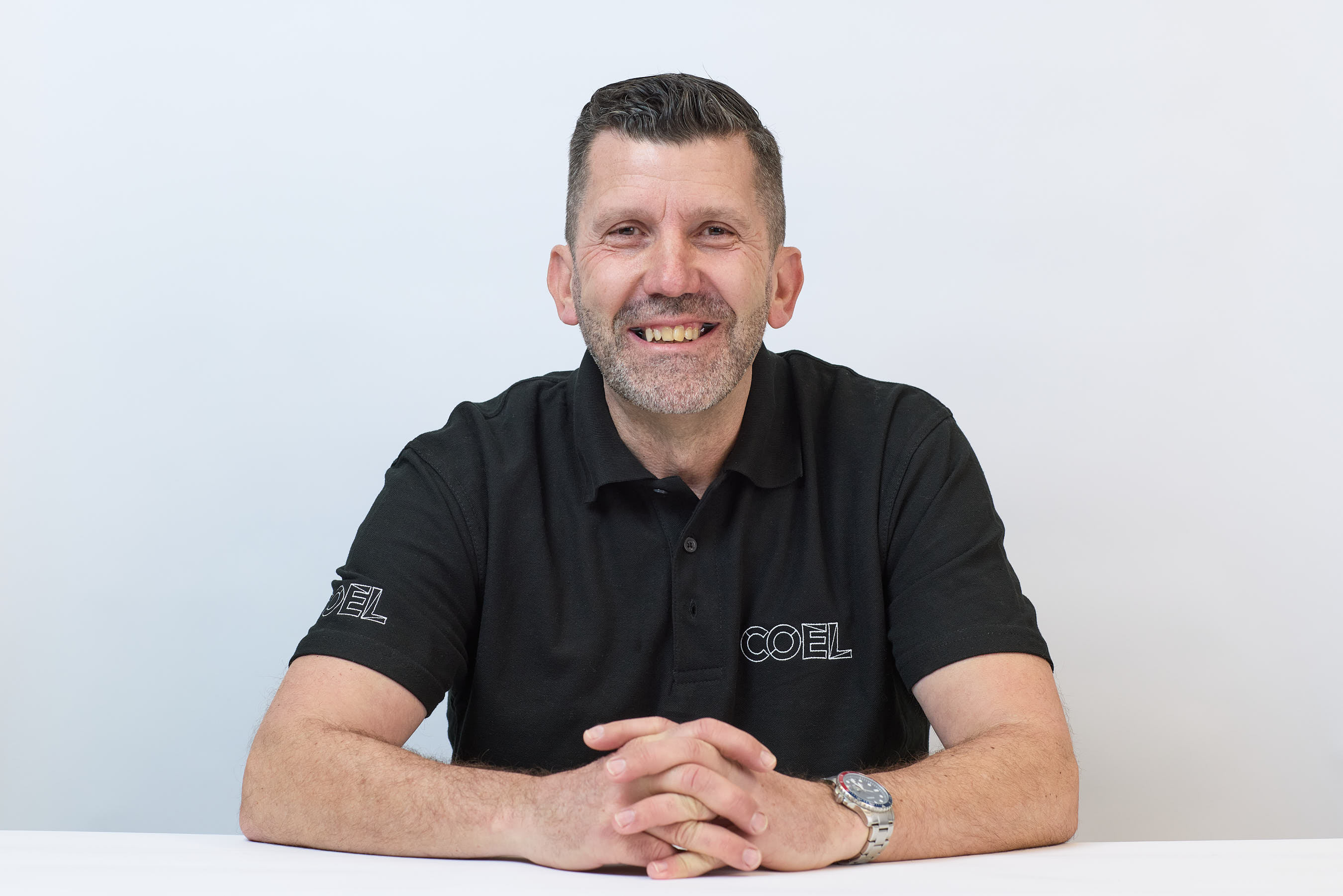Man smiling with arms crossed, wearing a black COEL-branded polo shirt against a plain white background.