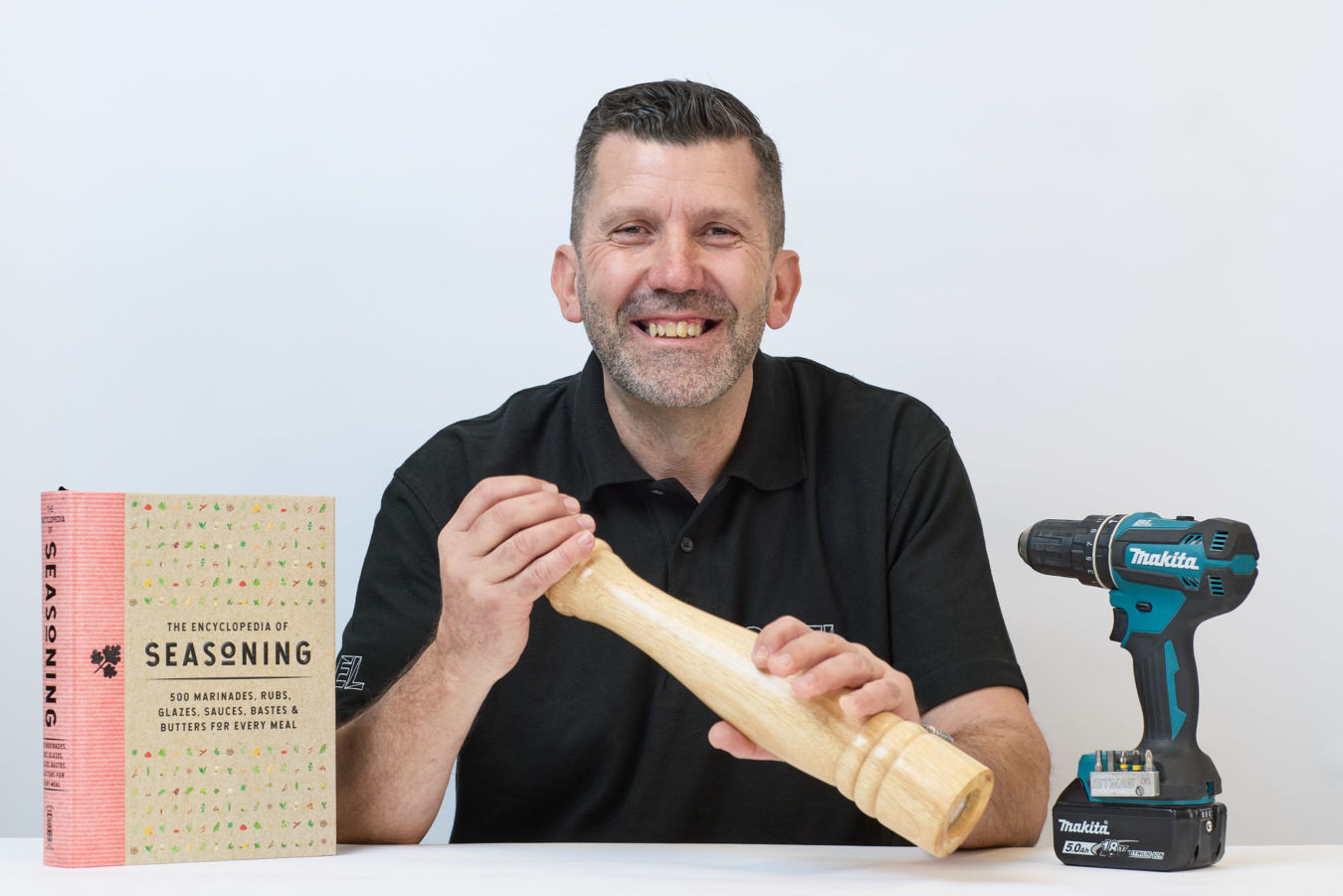 Individual holding a large wooden pepper mill, smiling, with a "Makita" drill and "The Encyclopedia of Seasoning" book beside them on a white table.