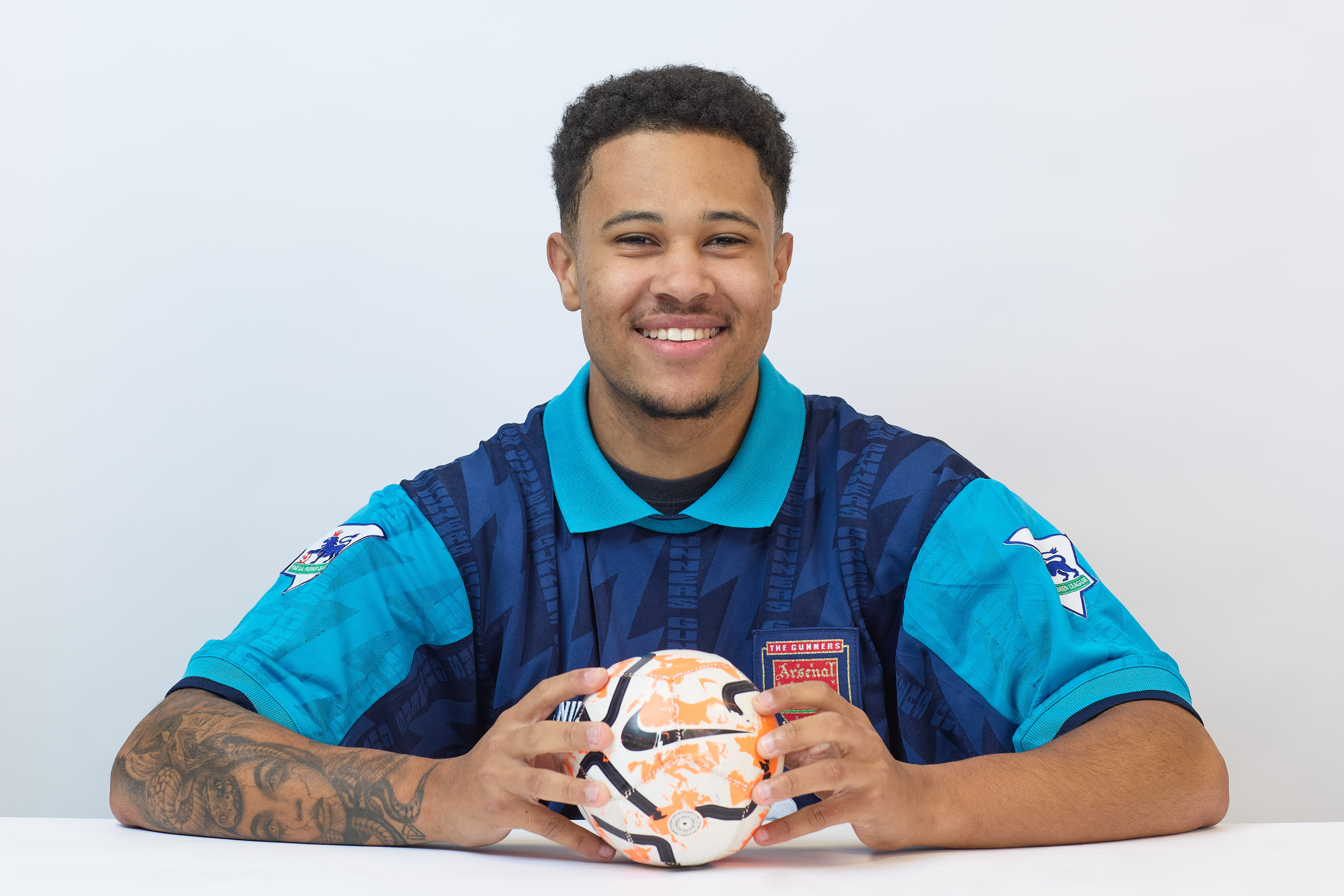 A smiling person holds a colorful soccer ball, wearing a blue vintage Arsenal soccer jersey, seated against a plain white background.