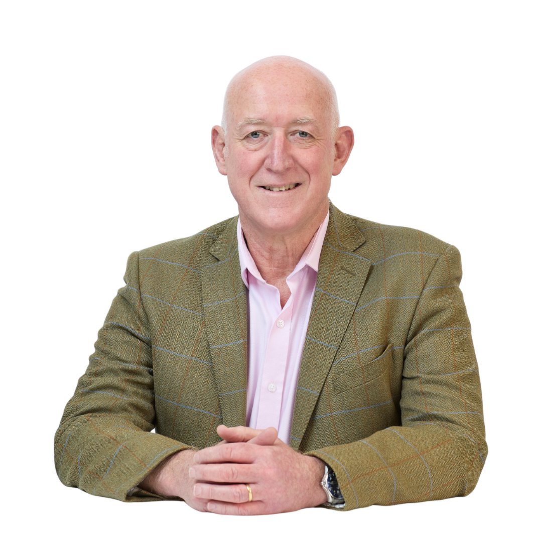 A man wearing a brown plaid suit jacket and a pink shirt sits with hands clasped, smiling, against a plain white background.