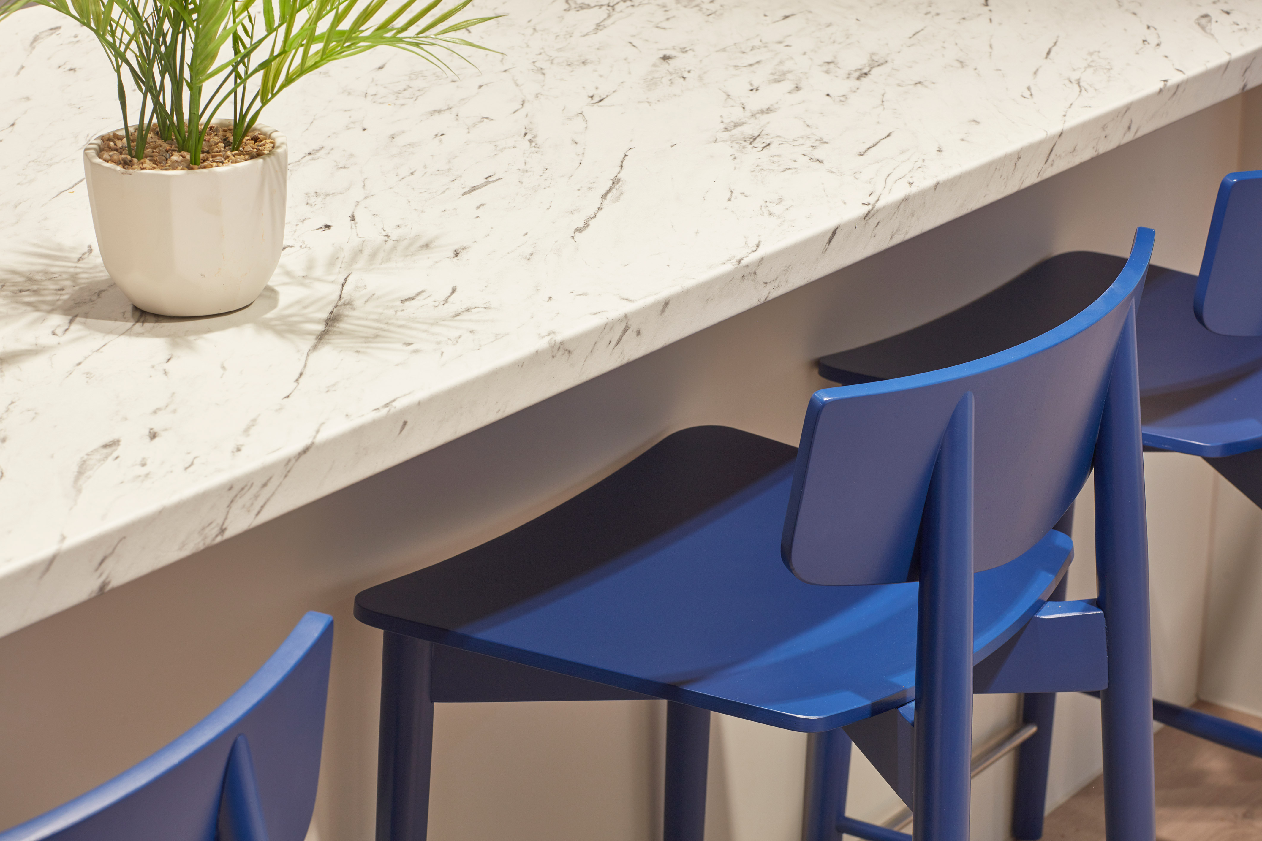 Blue bar stools line a white marble countertop, with a small potted plant on the surface, in a modern kitchen setting.