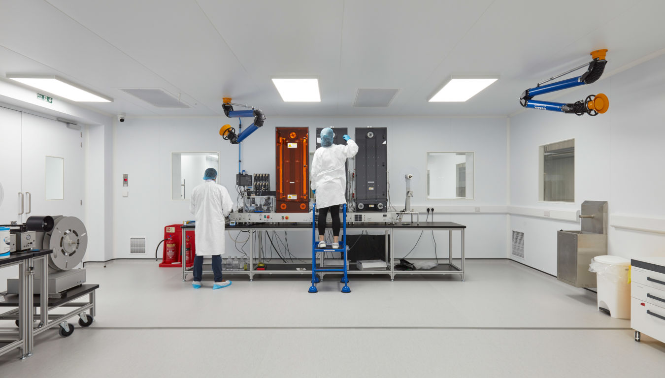 Cleanroom laboratory with two technicians in protective clothing working under bright lighting, surrounded by lab equipment and overhead exhaust systems.