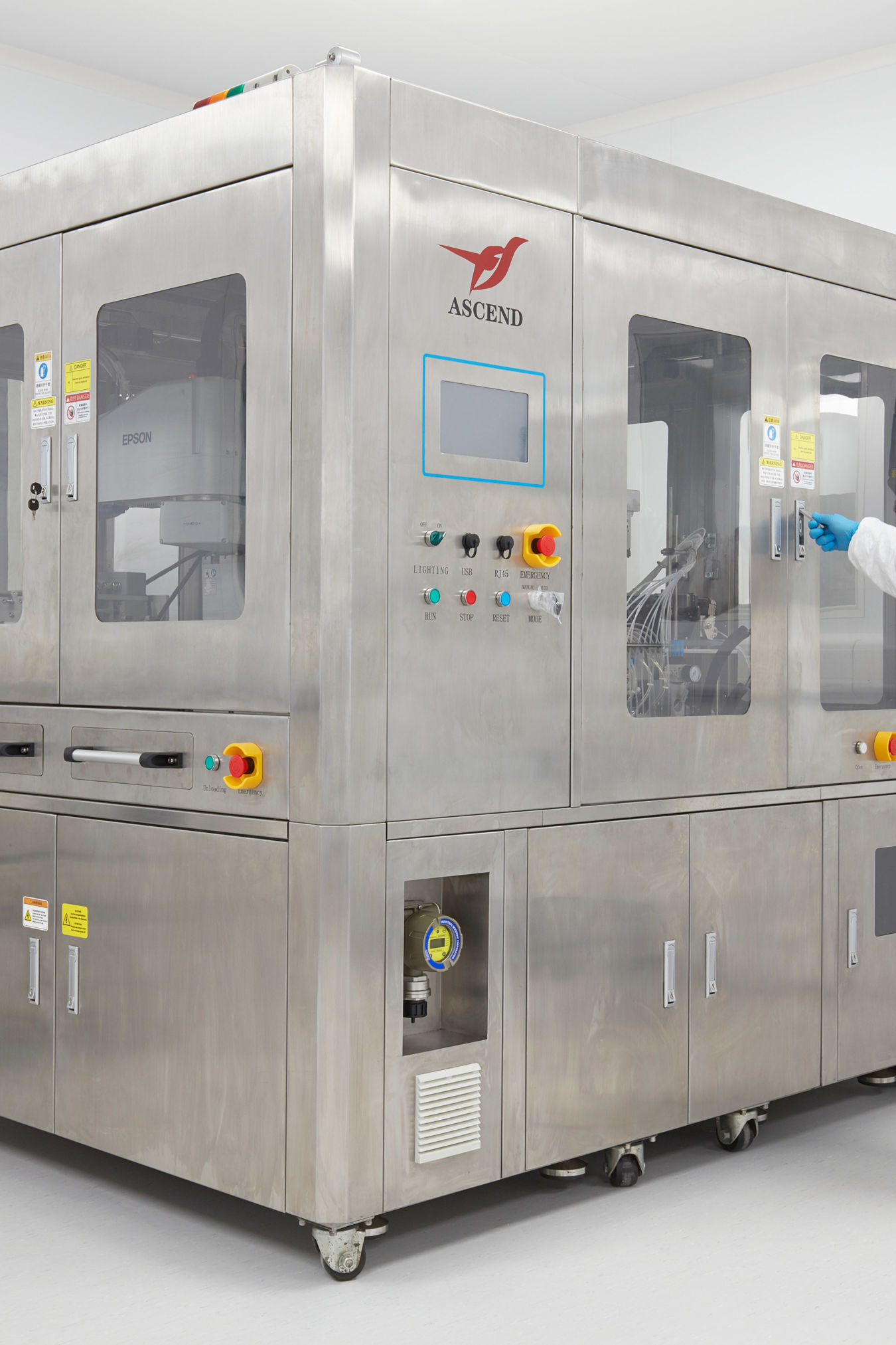Laboratory technician operating a large stainless steel automated machine inside a cleanroom, wearing protective clothing, gloves, and a hairnet.