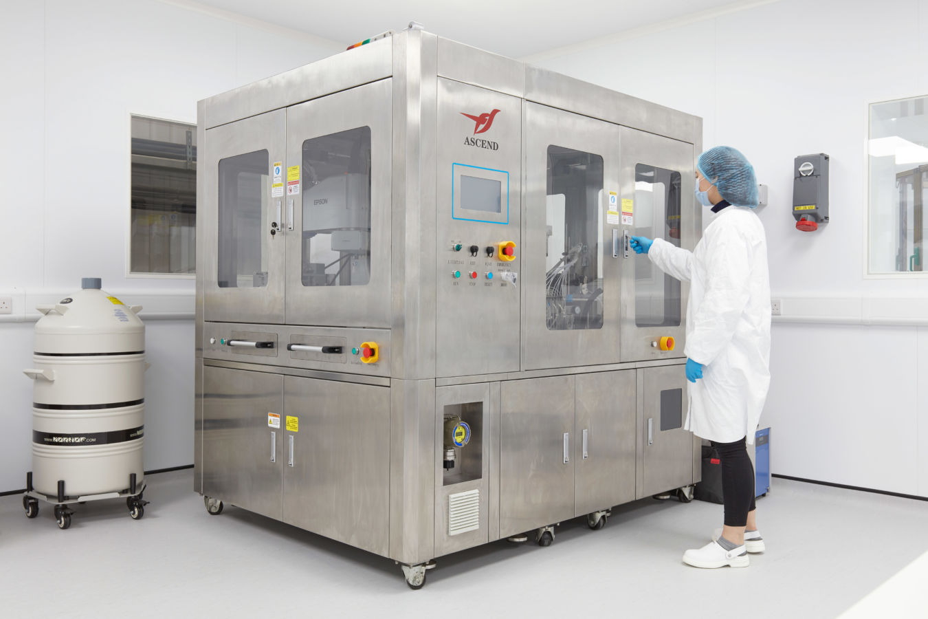 Laboratory technician operating a large stainless steel automated machine inside a cleanroom, wearing protective clothing, gloves, and a hairnet.