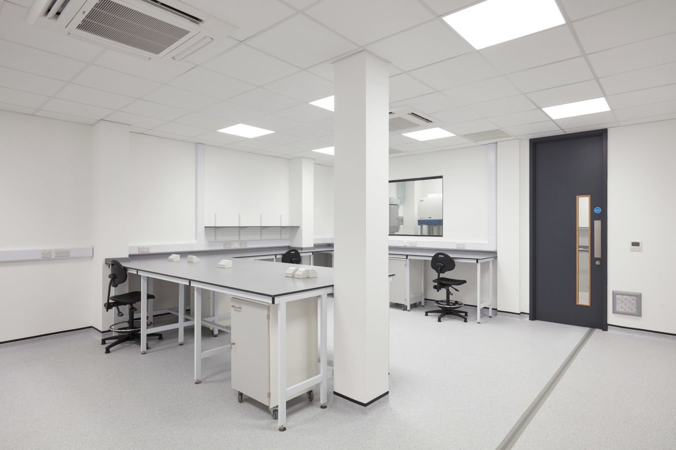 Clean, modern laboratory space with white walls, workbenches, rolling drawers, lab stools, and a bright ceiling light grid.
