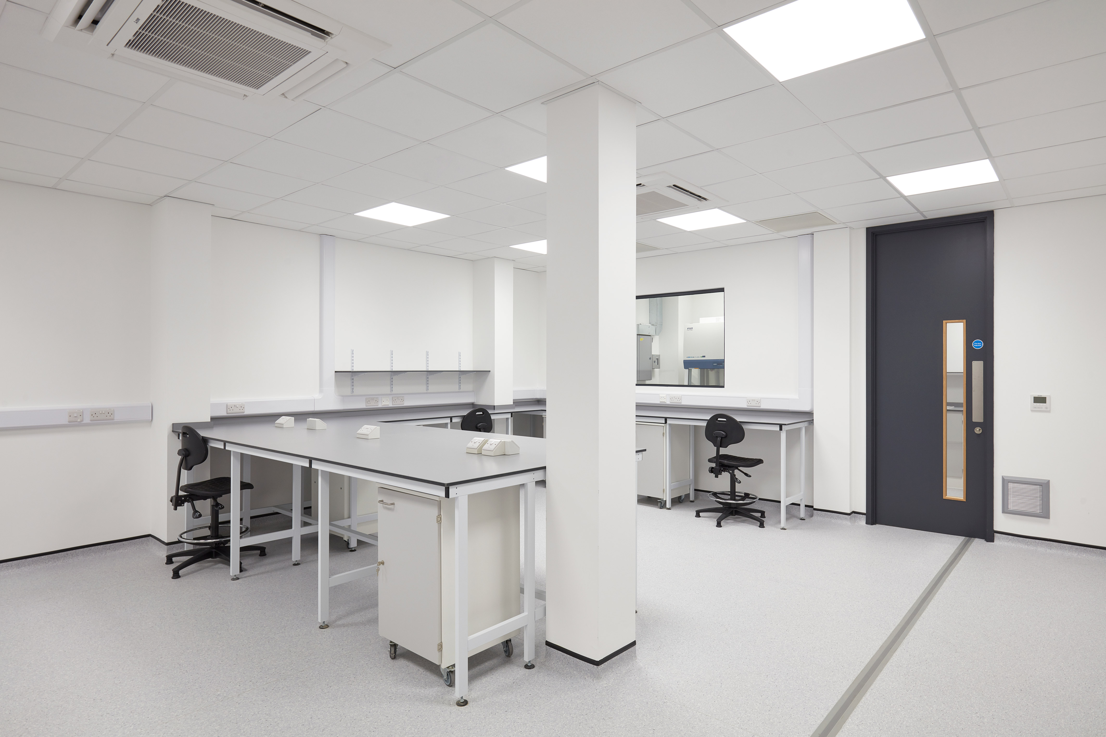 Clean, modern laboratory space with white walls, workbenches, rolling drawers, lab stools, and a bright ceiling light grid.