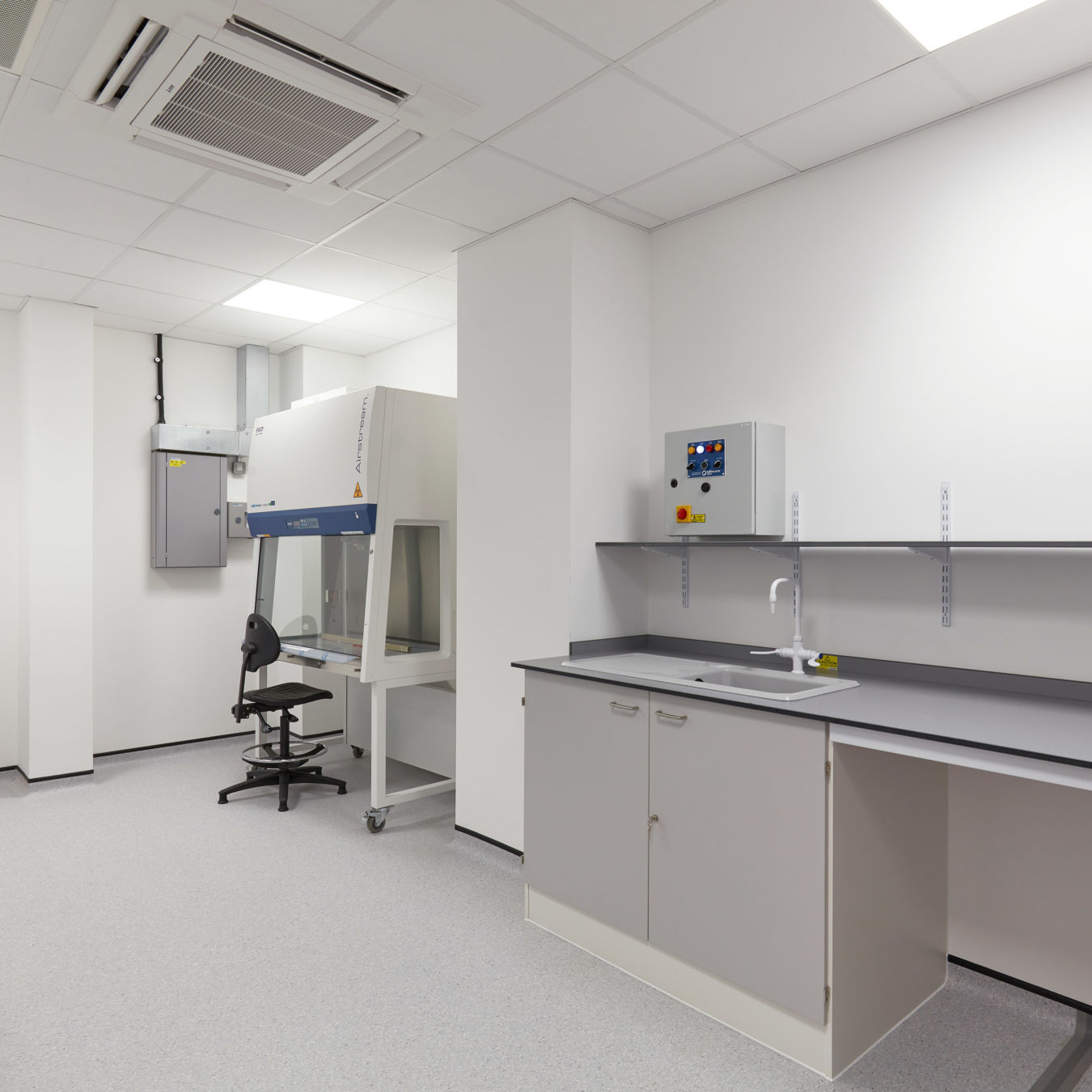 Sterile laboratory space with clean white walls, grey cabinetry, a fume hood workstation, overhead lighting, and a ventilation system