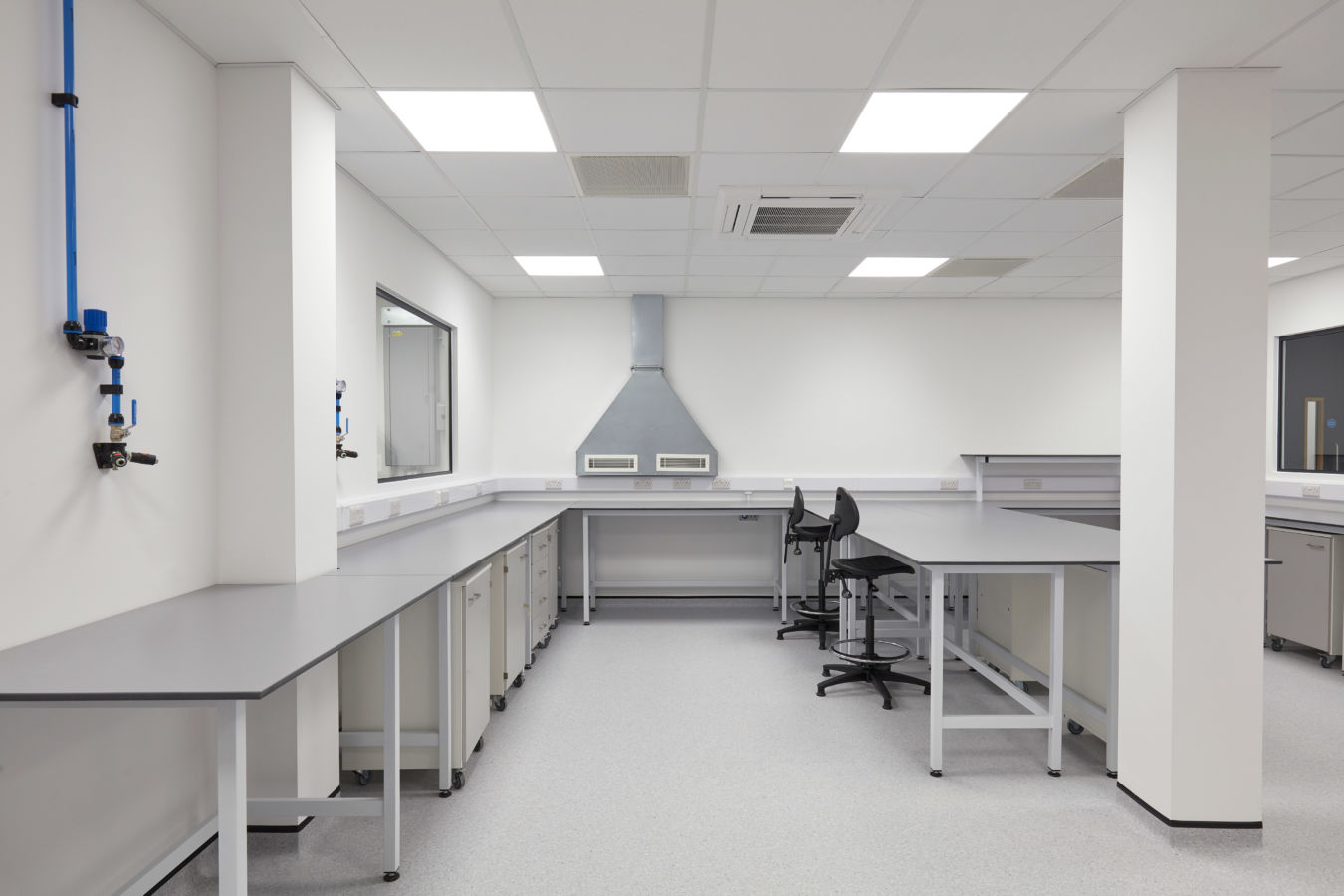 Clean and modern laboratory room with white walls, long countertops, overhead ventilation hood, office chairs, and bright ceiling lights