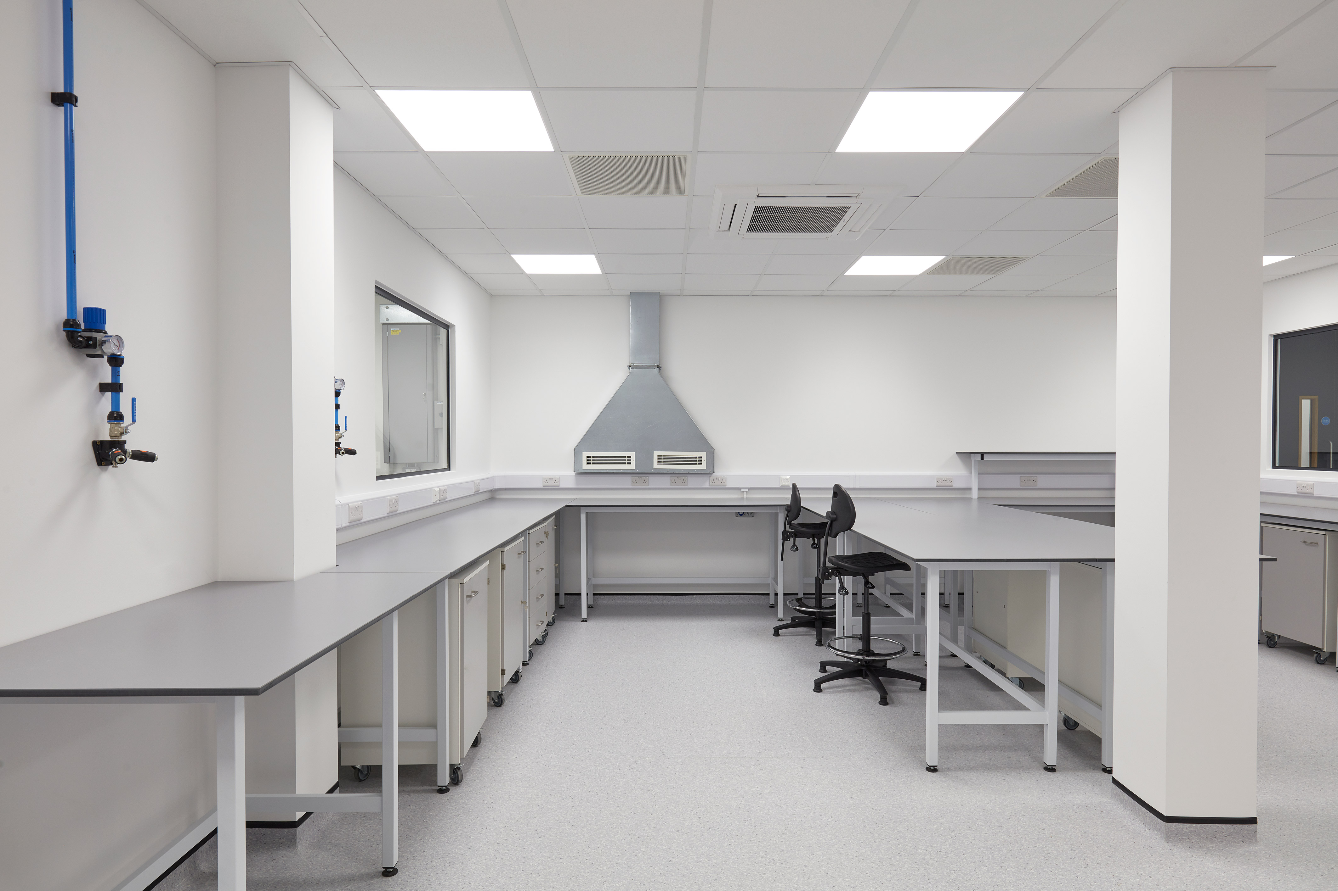 Clean and modern laboratory room with white walls, long countertops, overhead ventilation hood, office chairs, and bright ceiling lights