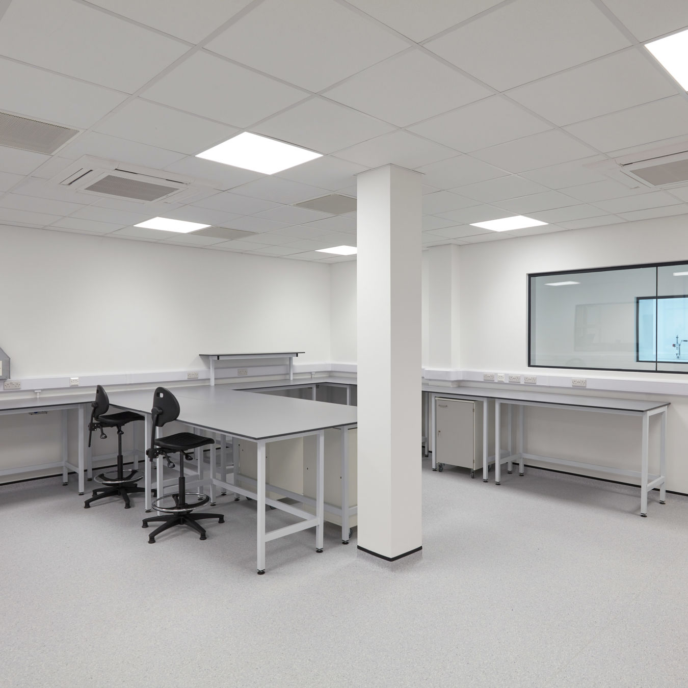 Spacious, white laboratory with multiple workbenches, lab stools, overhead lighting, and a large interior observation window